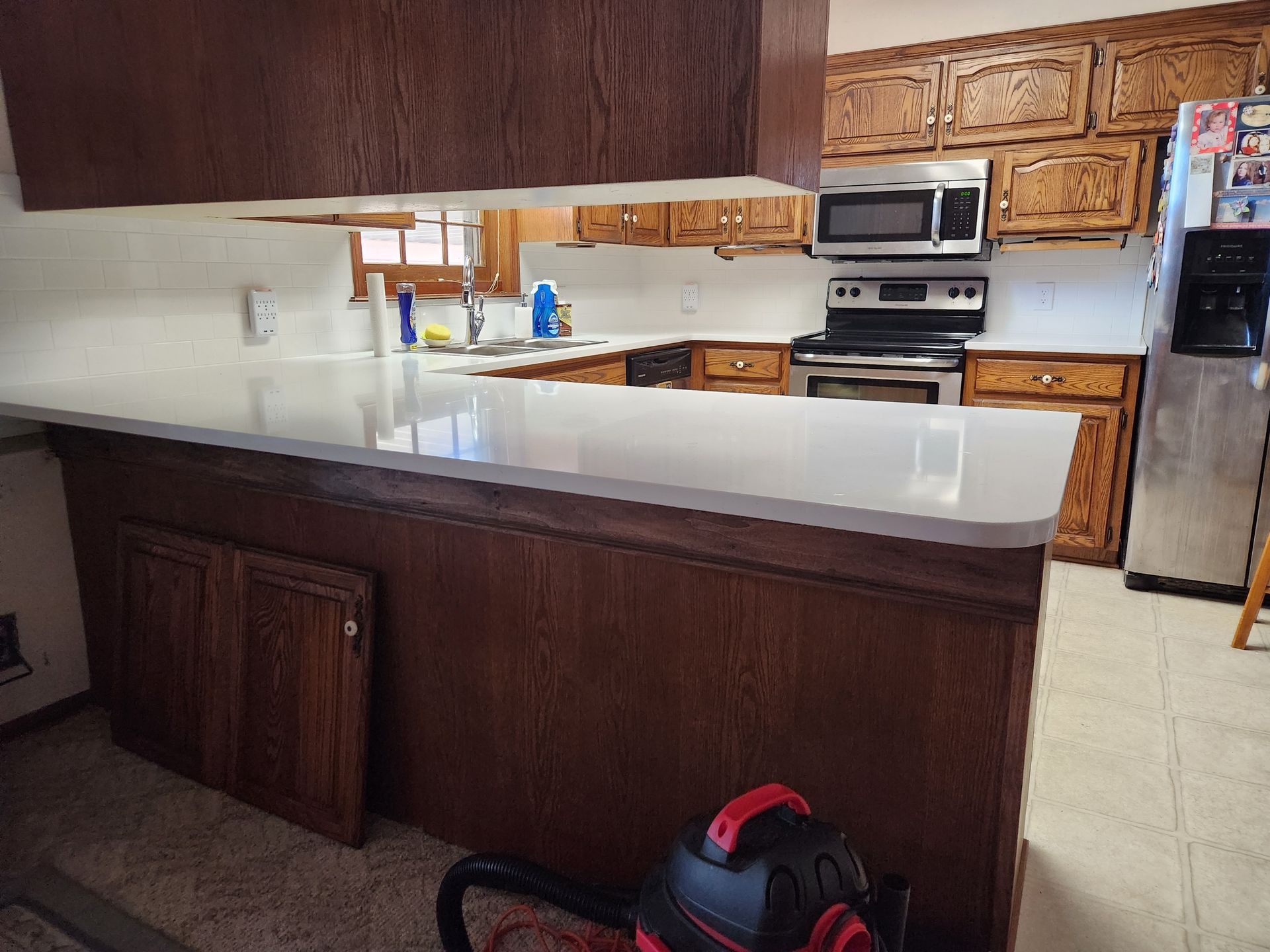 Kitchen with light countertops, dark wood cabinets, and stainless steel appliances. A vacuum cleaner sits in front.