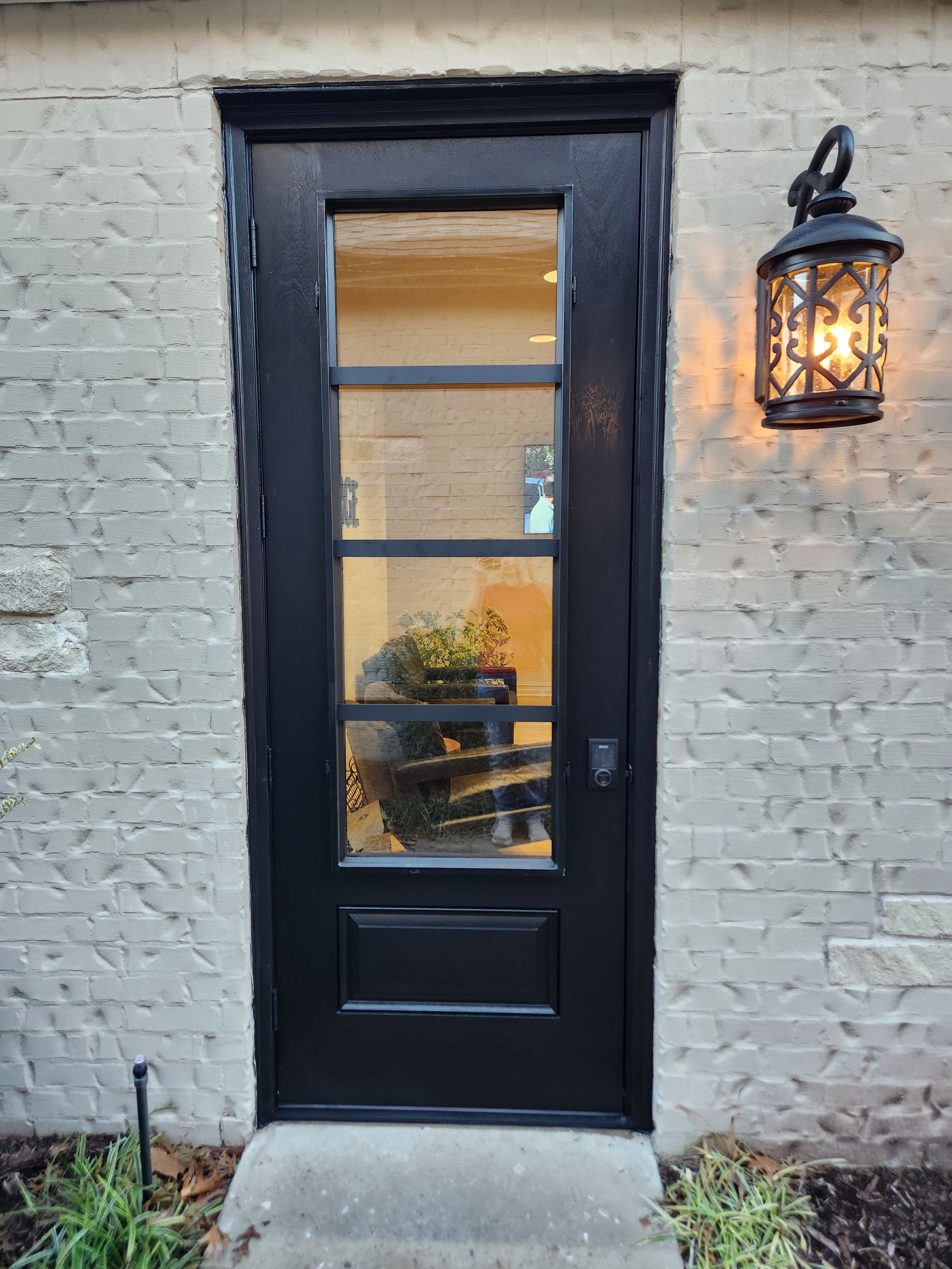 Black door with rectangular glass panels, mounted in a light-colored brick wall with decorative lantern to the right.