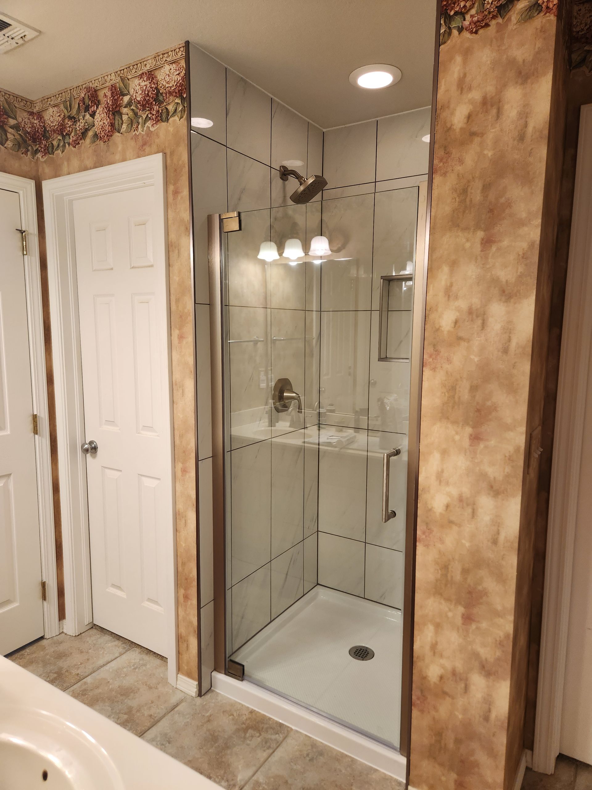 Shower with glass door, white walls, and a tiled floor inside a bathroom with floral wallpaper border.