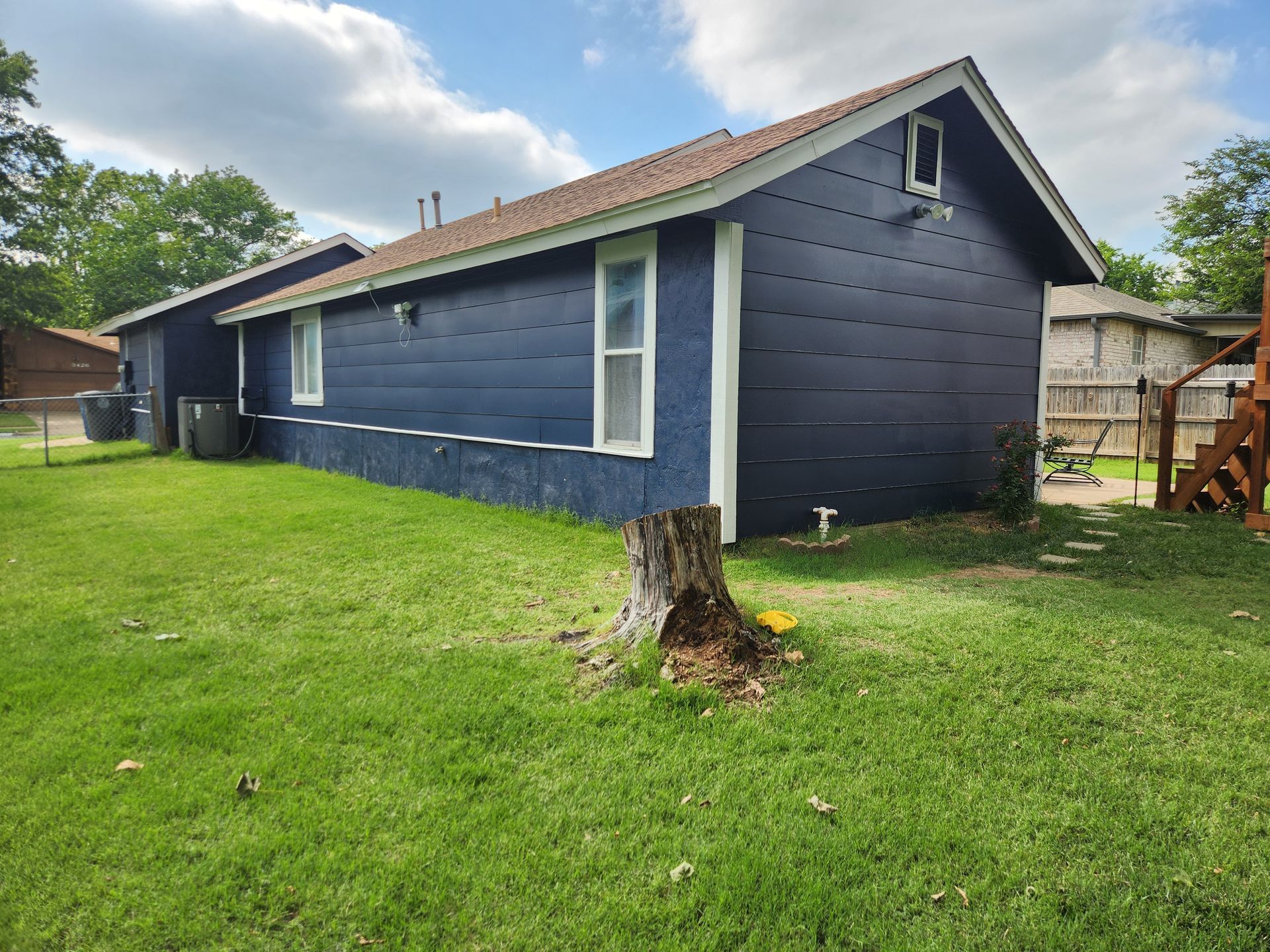 Backyard view of a dark blue house with white trim, green grass, a tree stump, and a wooden fence.
