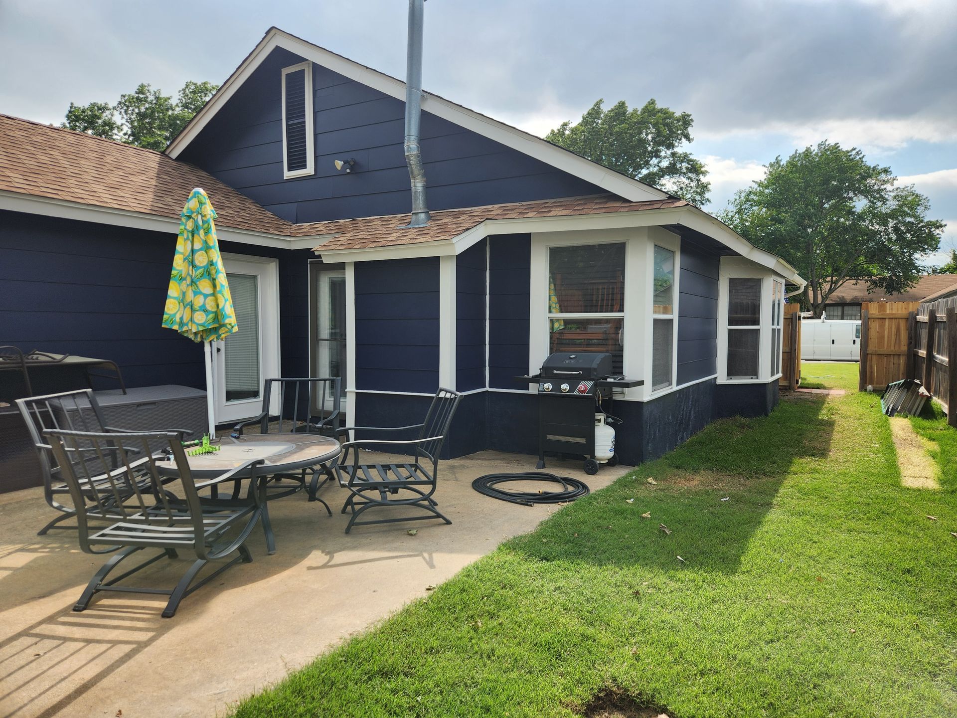 Blue house with patio furniture and grill on concrete, green lawn, sunny day.