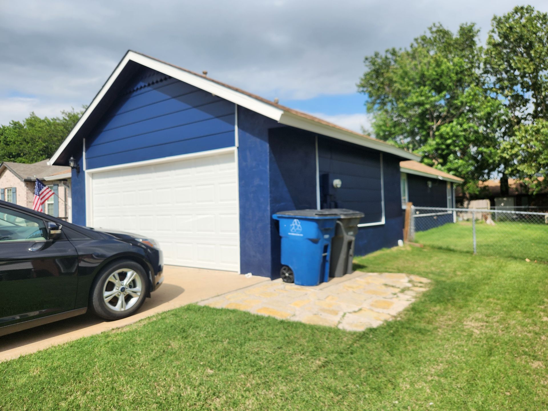 Blue house with white garage door, black car, and trash cans on a lawn.