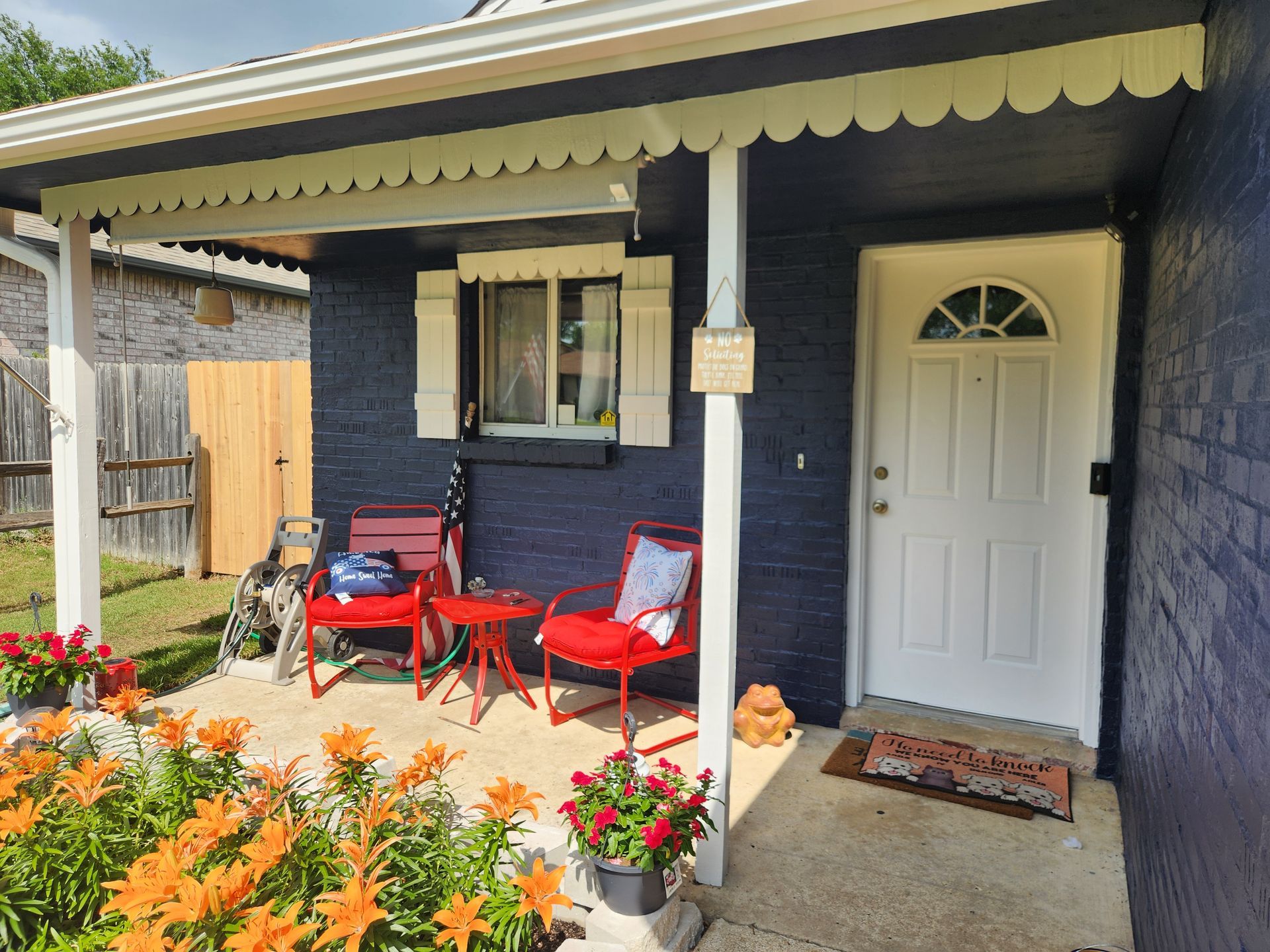 A blue house with a white door and porch. Red chairs and flowers are in front.