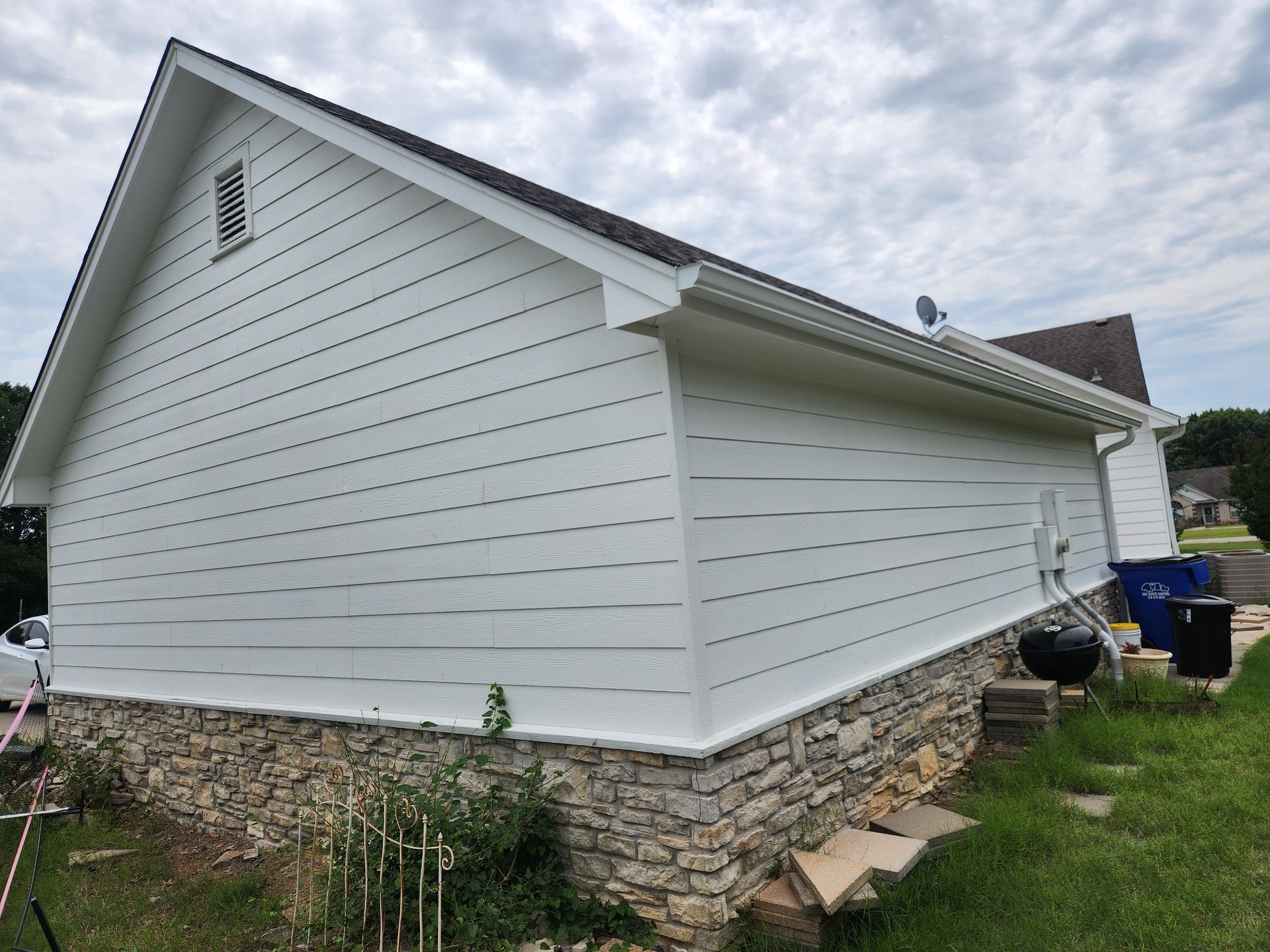 White siding on a house with a stone foundation, brown roof, and green lawn.