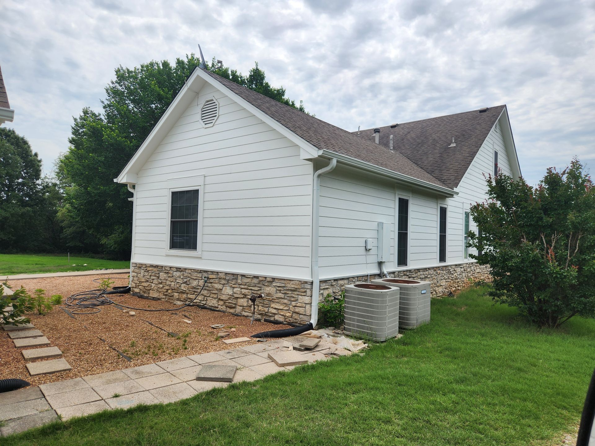 White house with stone base, dark roof, and green lawn under a cloudy sky.