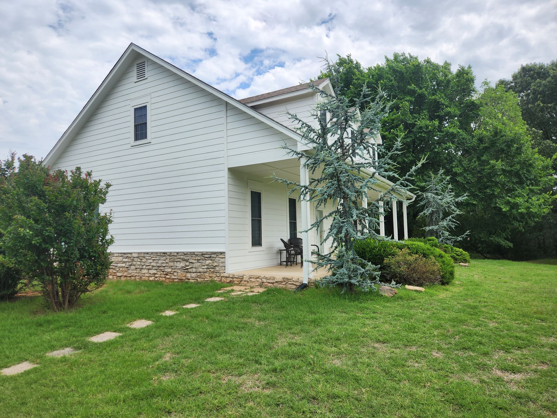 White farmhouse with stone foundation and porch, set in a grassy yard under a cloudy sky.