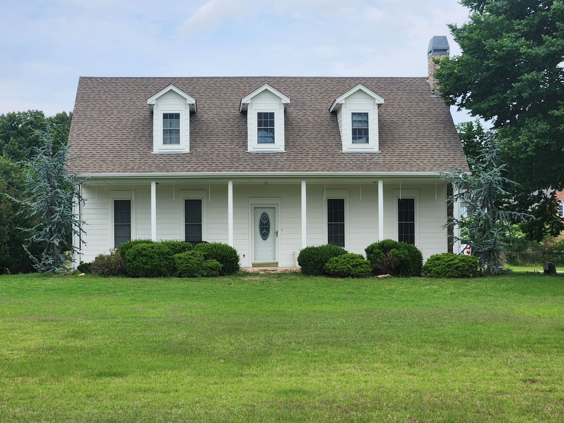 White house with a brown roof, three dormer windows, a porch, and green lawn.