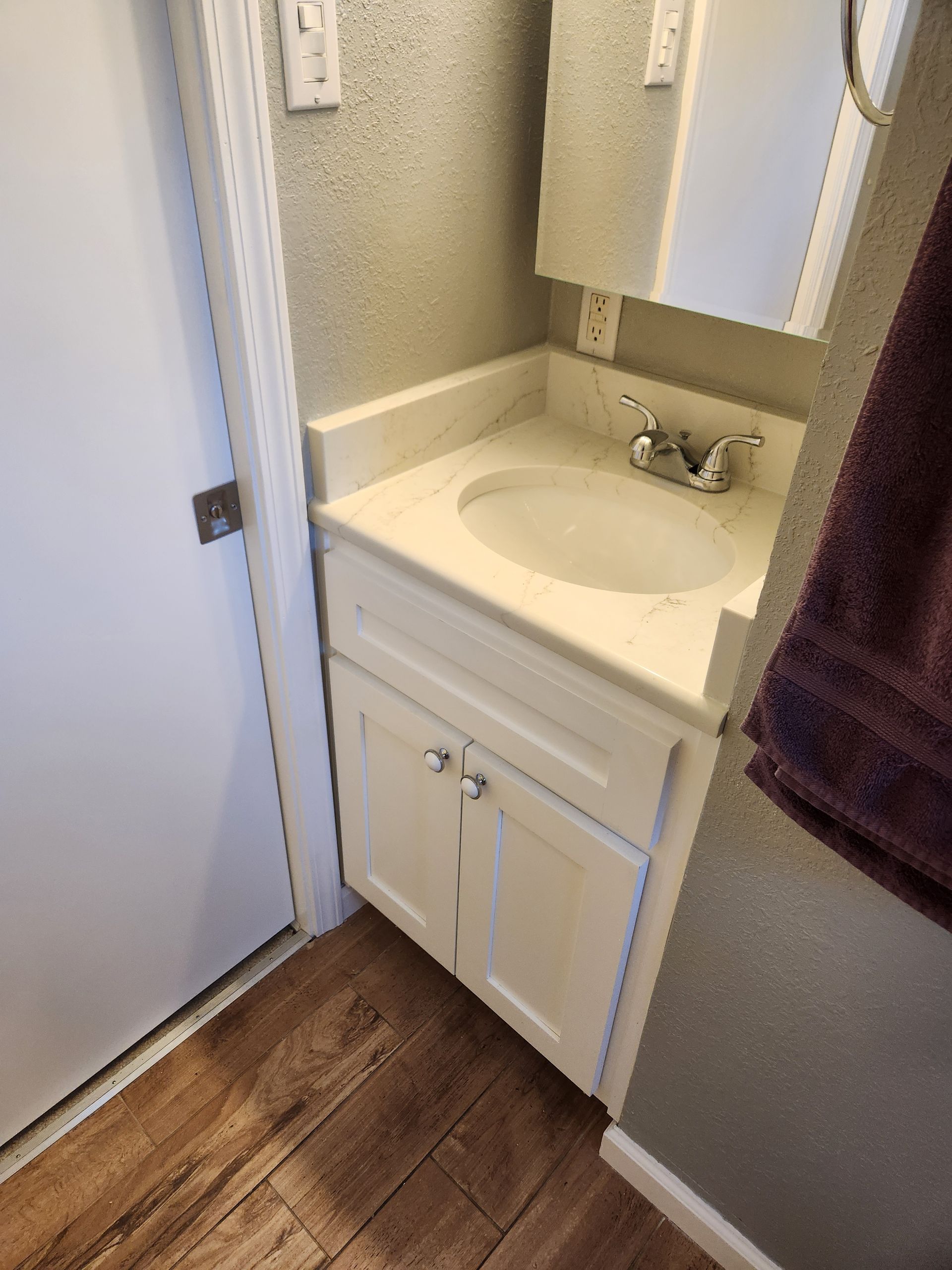 Small bathroom with white vanity, sink, and mirror. Door to the left, towel on the right.