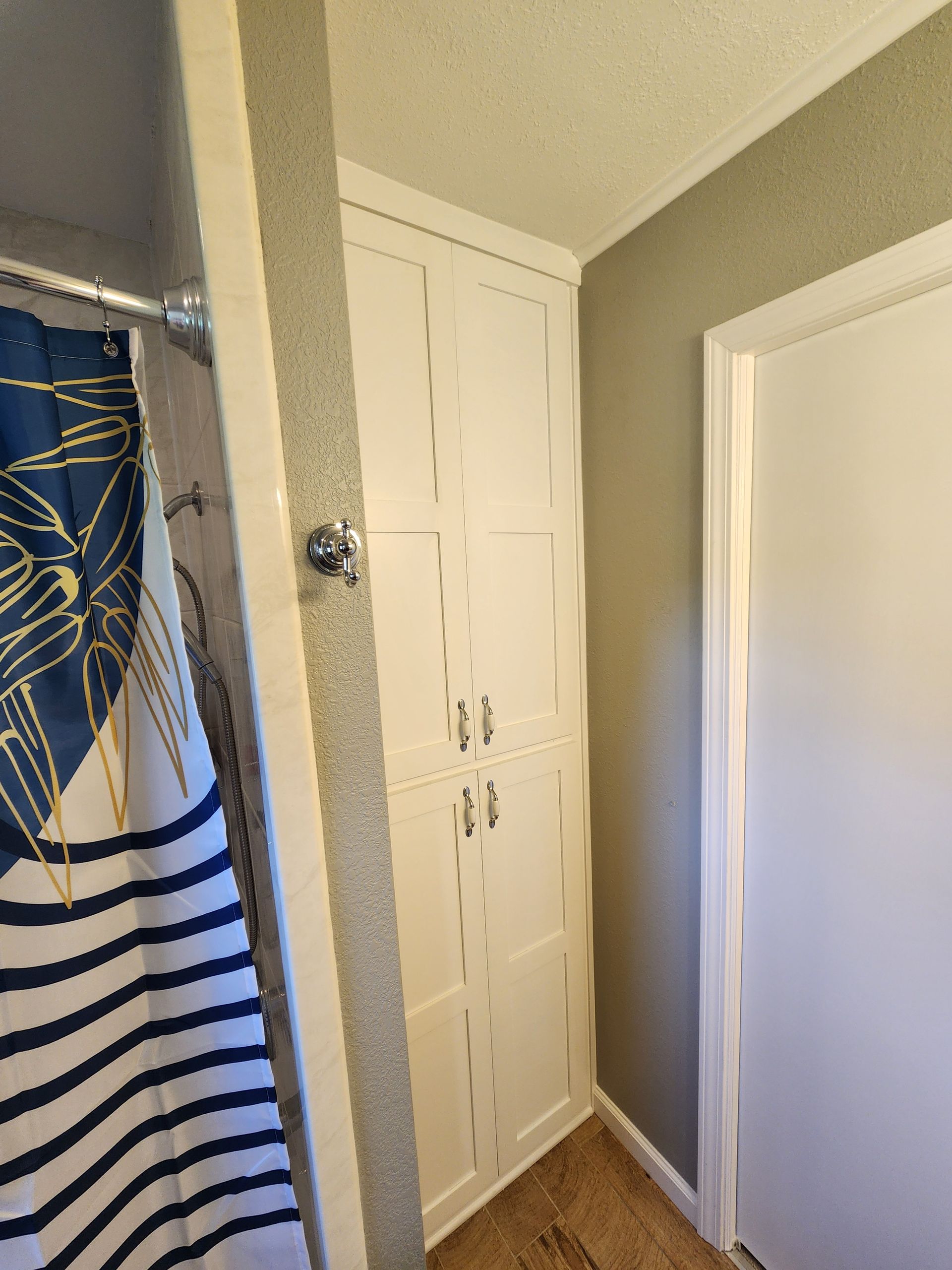 Tall white cabinet built into a corner, next to a shower with a blue and white striped curtain and a white door.