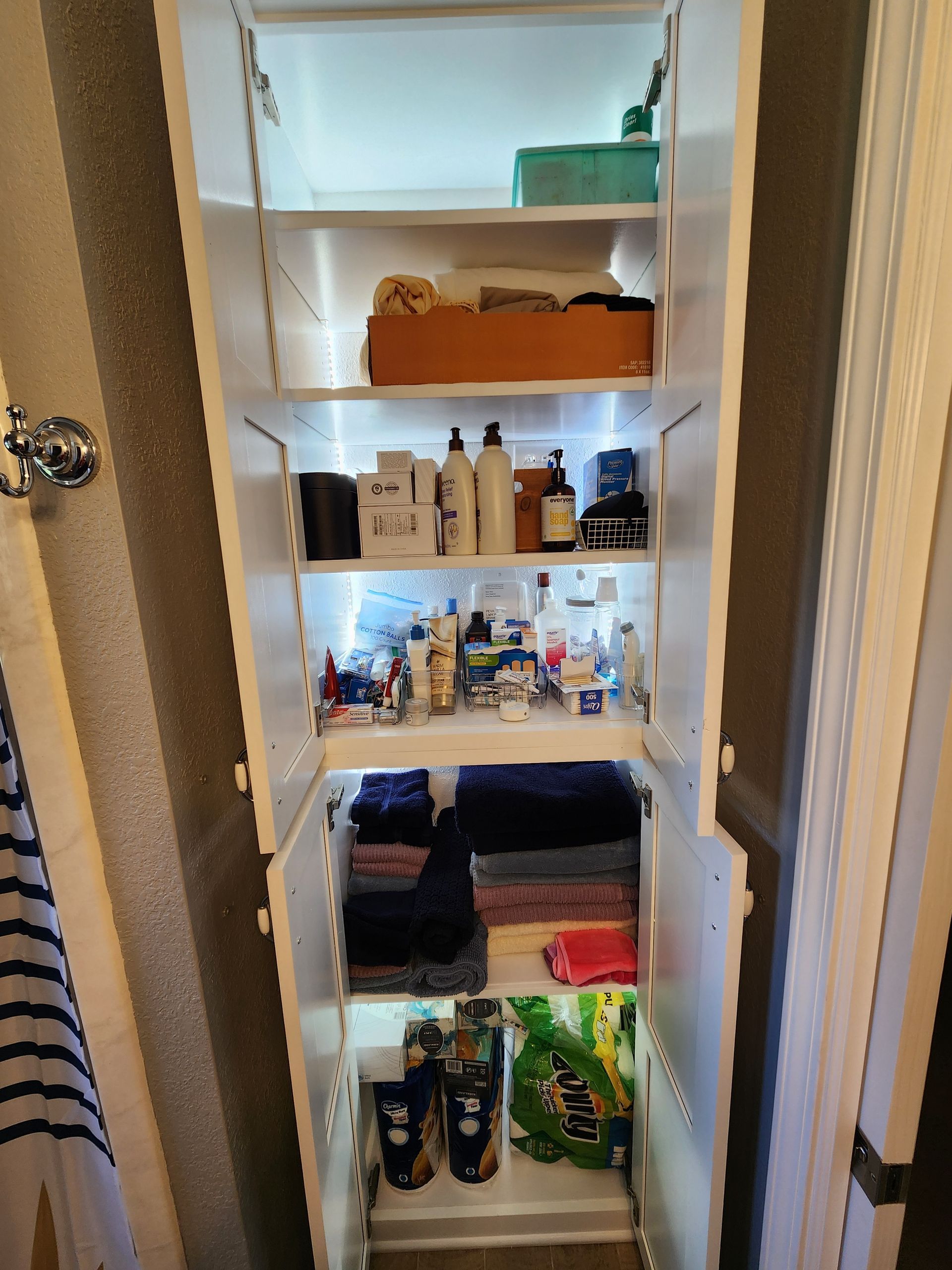 Tall, open white bathroom cabinet with shelves.  Holds towels, toiletries, and cleaning supplies. Interior has LED lighting.