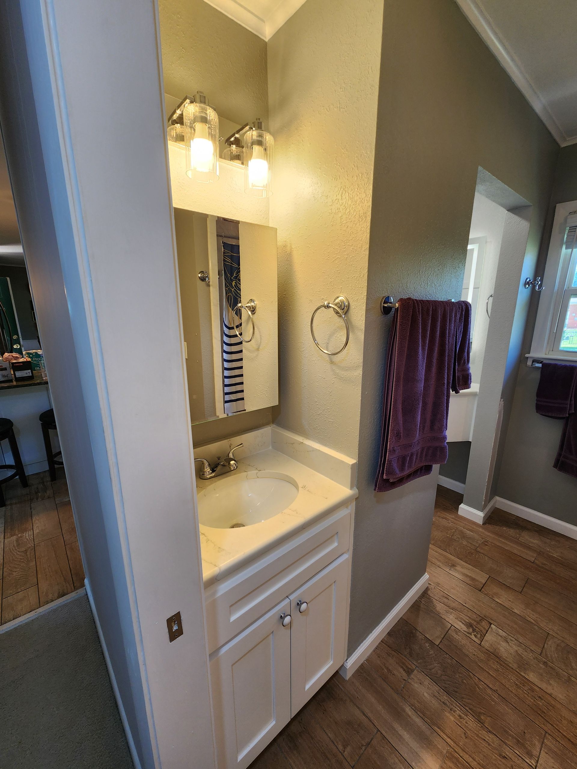 Small bathroom with white vanity, mirror, and towel rack. Gray wall, wooden floor, and a glimpse into another room.