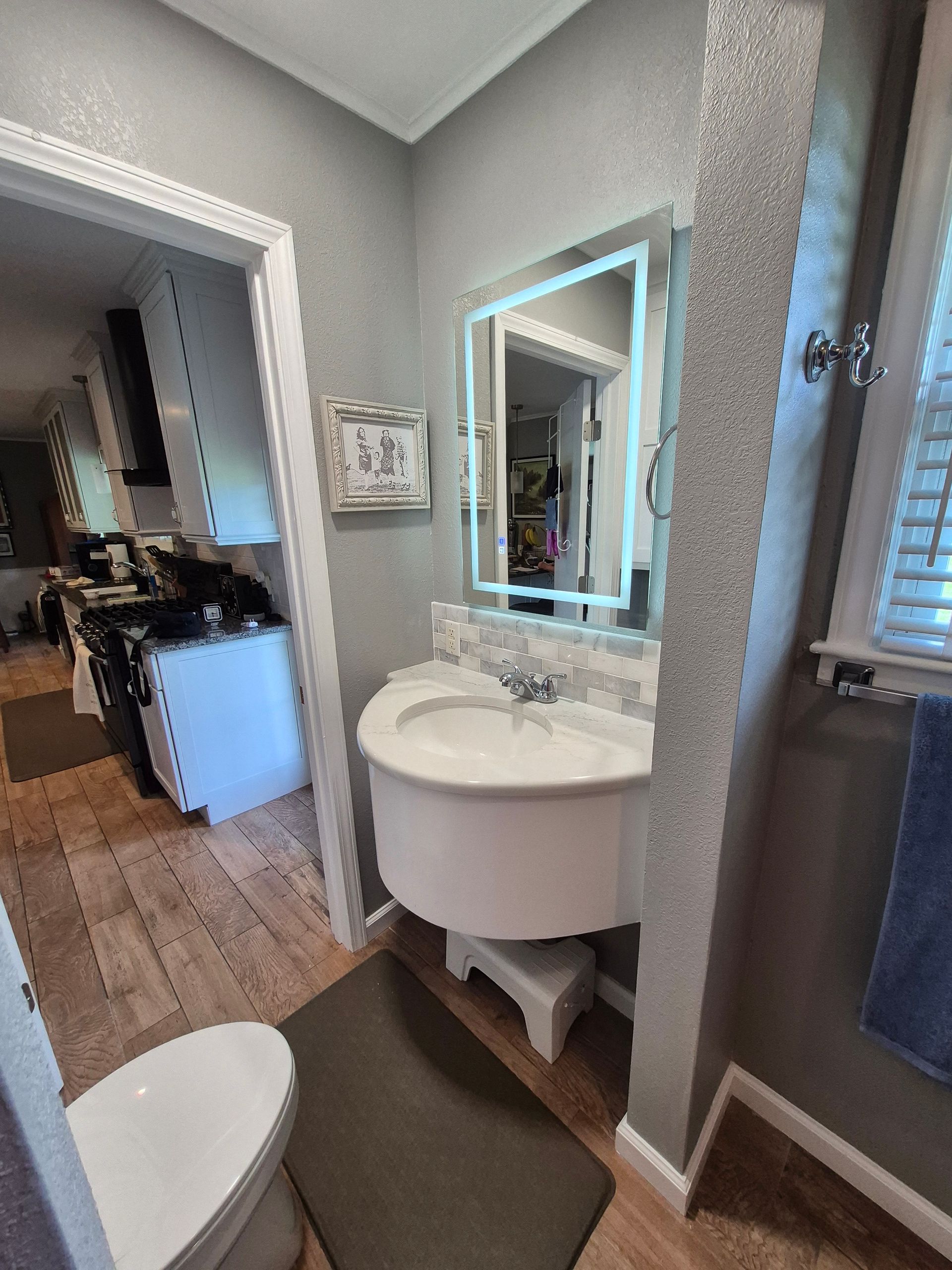 Bathroom with white pedestal sink, lit mirror, gray walls, and open doorway to a kitchen.
