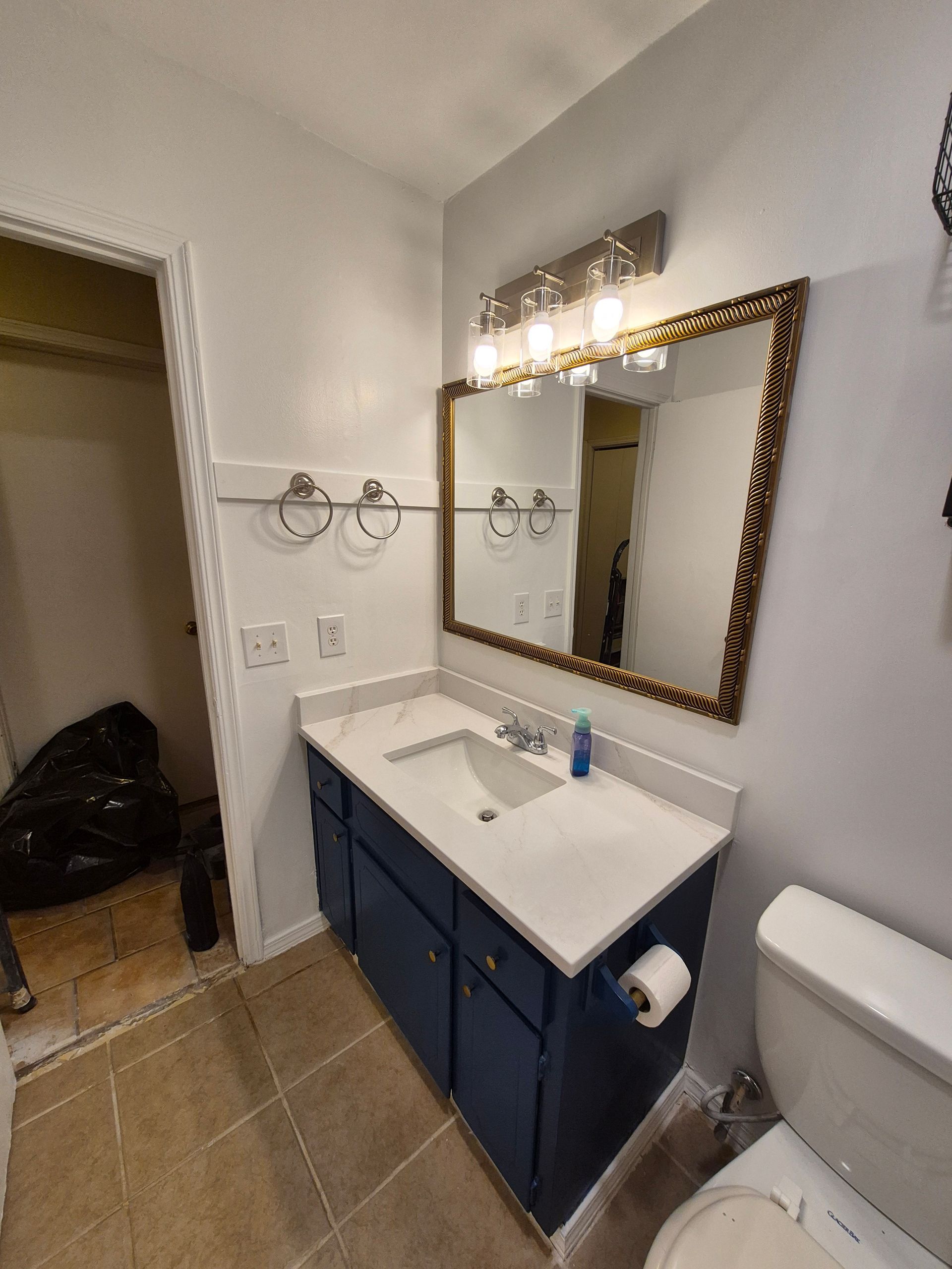 Bathroom with blue vanity, gold-framed mirror, and silver towel rings on a light gray wall.