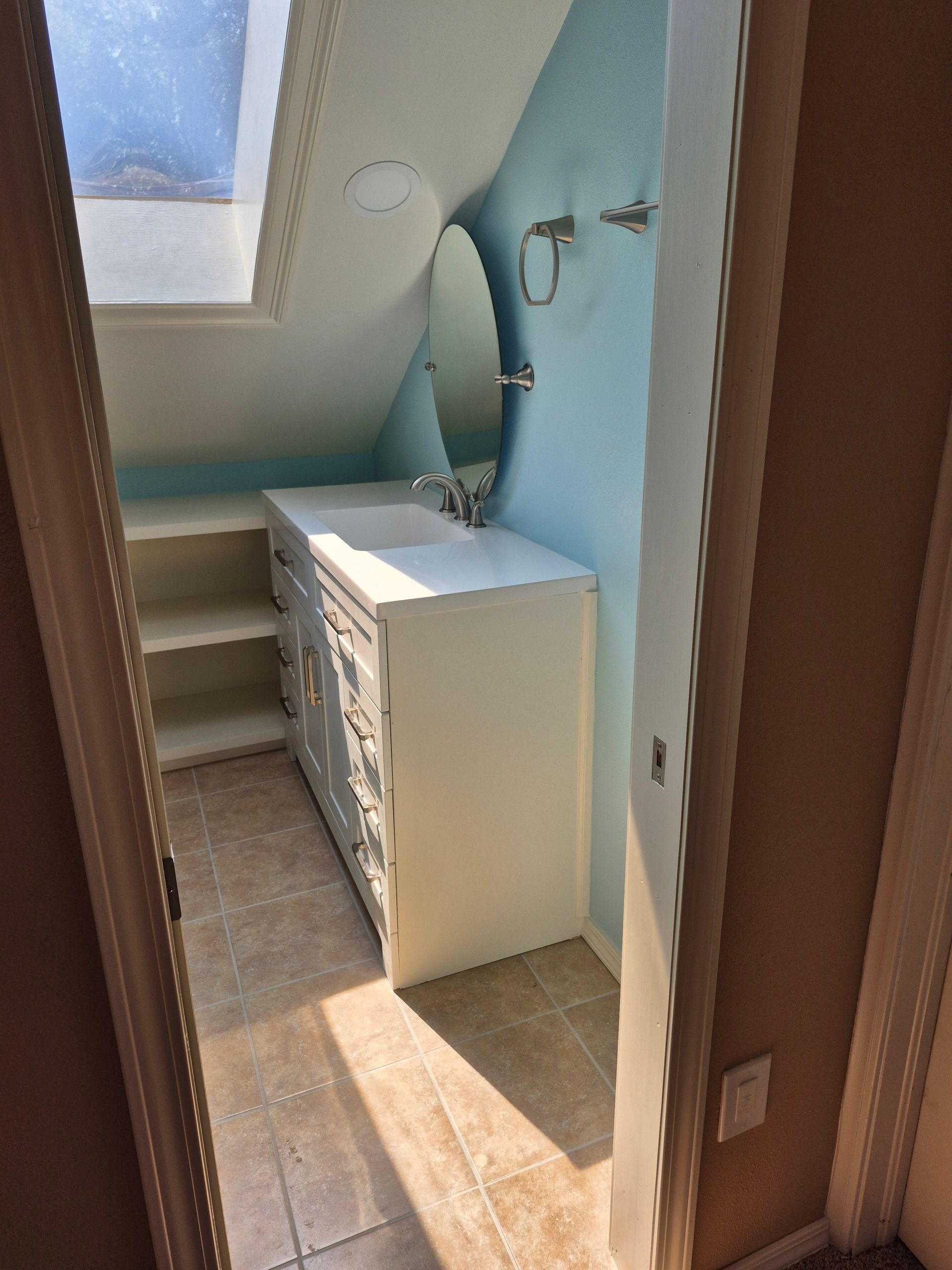 Bathroom with white vanity, round mirror, and tiled floor, under a sloped ceiling and window.