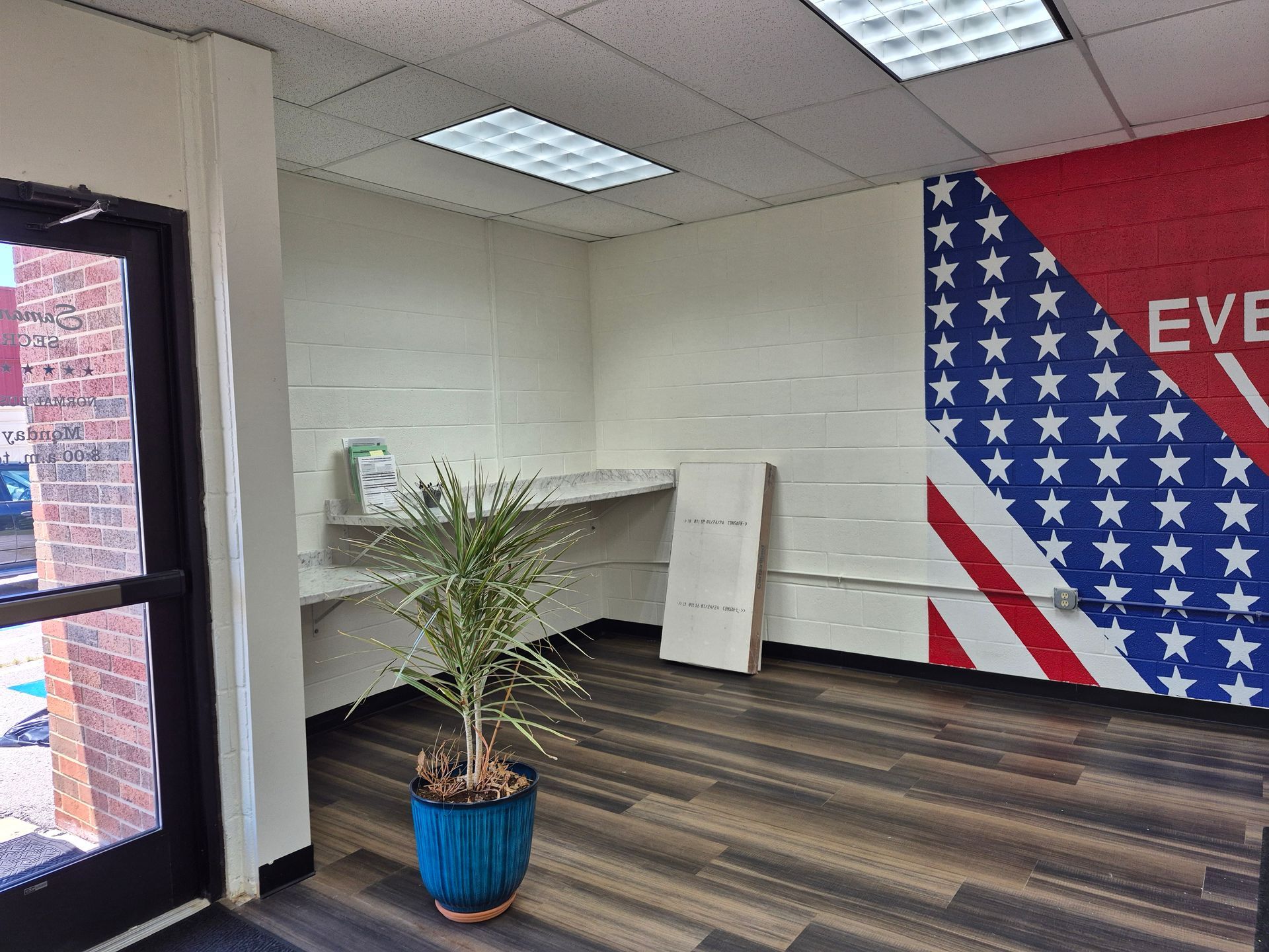 Interior view: Office with an American flag mural, a plant in a blue pot, and wood-like flooring.