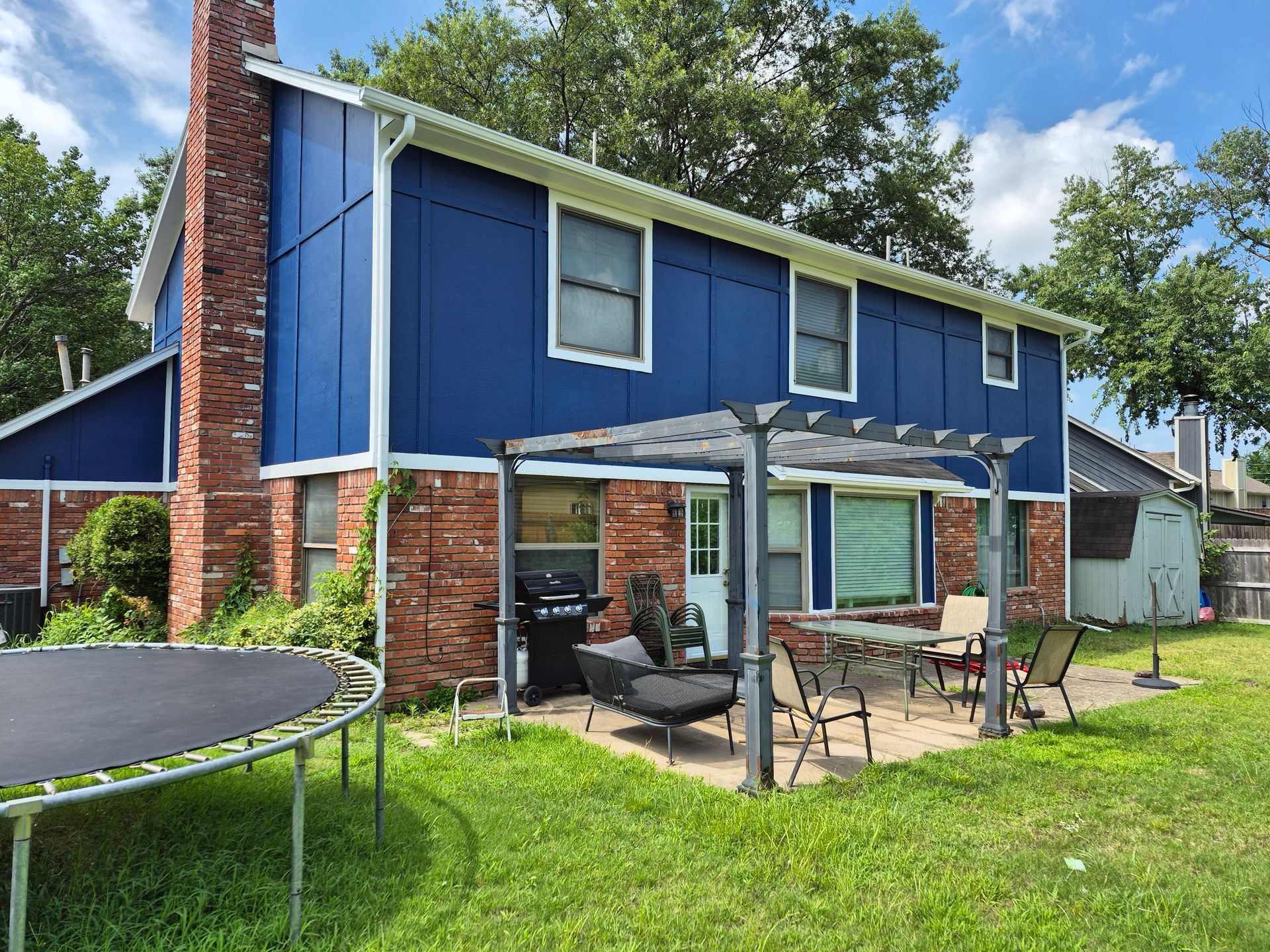 Backyard view of a house with blue siding, a red brick chimney, and a patio with a pergola and outdoor furniture.