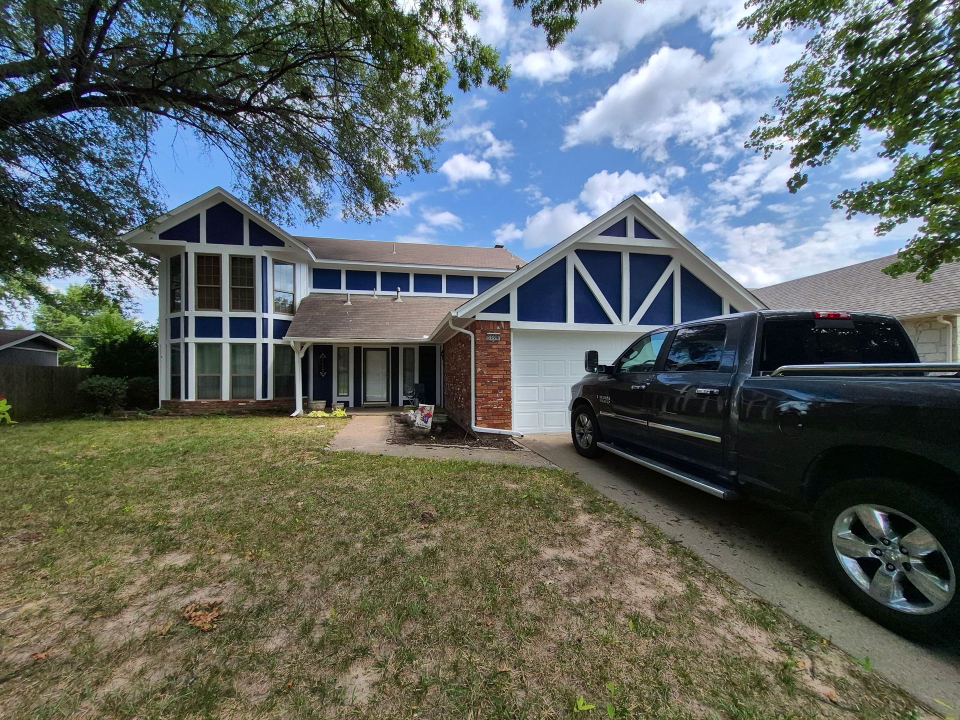 Two-story house with blue trim, brick accents, and a black pickup truck in the driveway. Green lawn, blue sky.