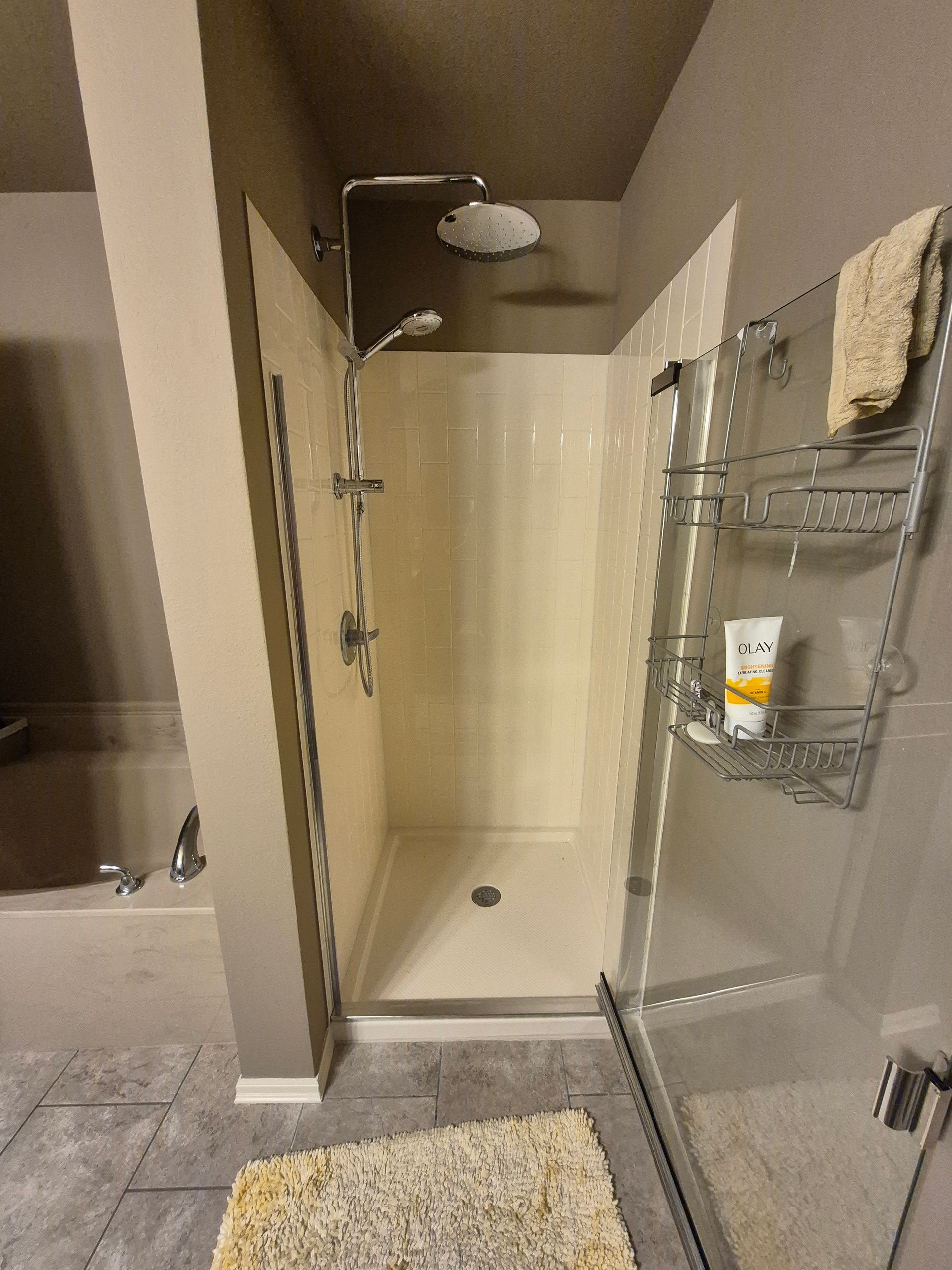 Shower stall with glass door, beige walls, chrome fixtures. A shelf and towel hang on the door.