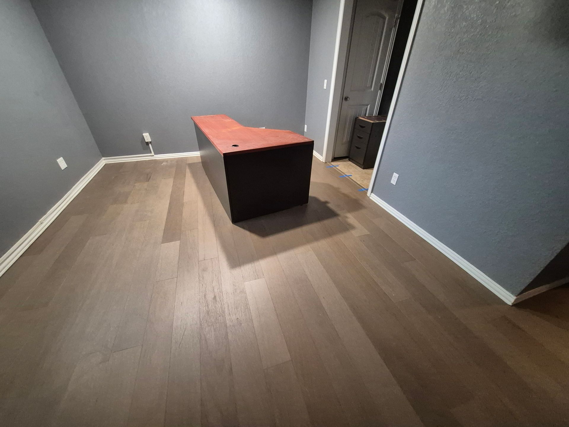 Empty room with wood floors, gray walls, and a large brown desk. A door is visible in the distance.