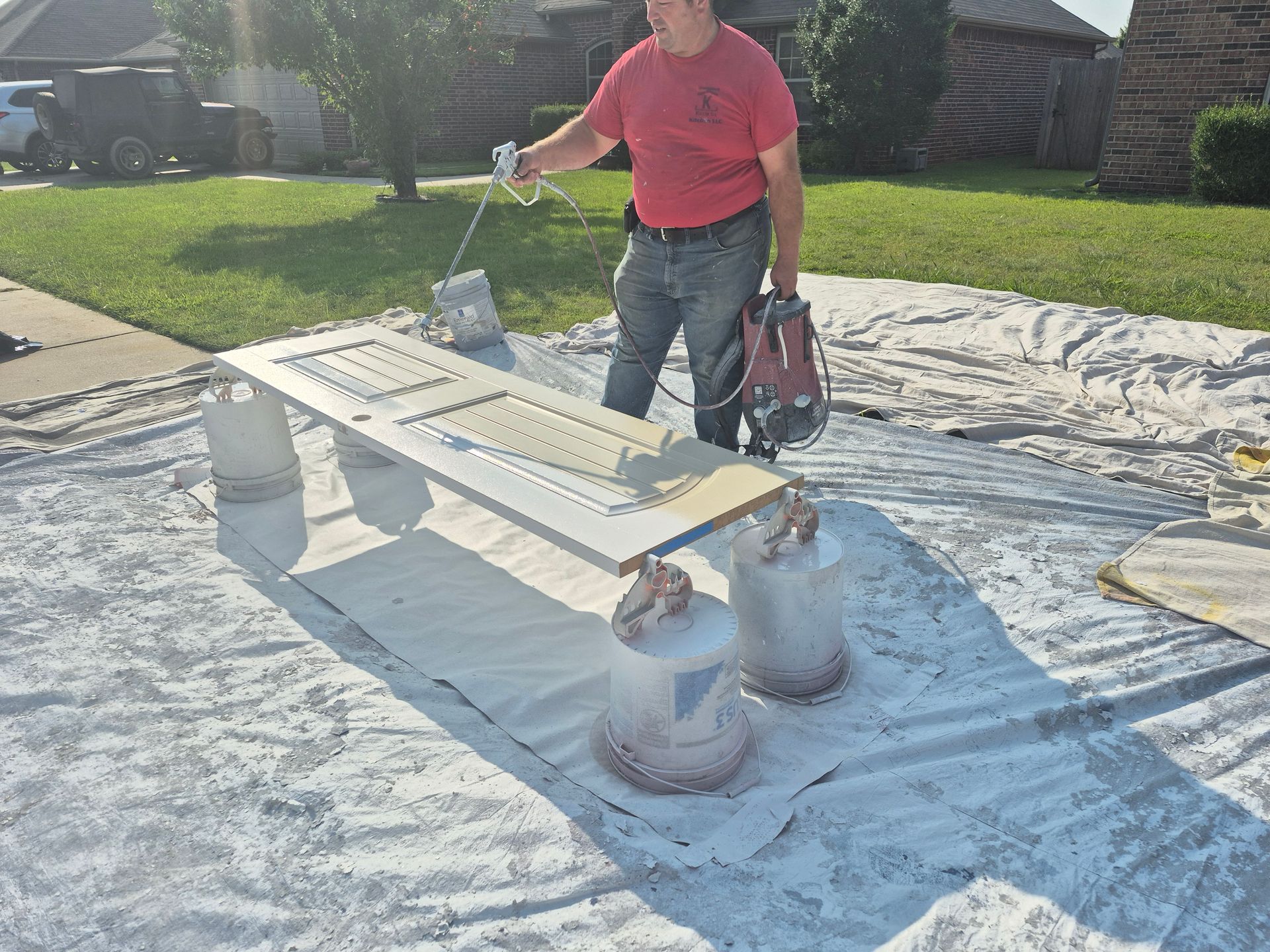 Person spray painting a white door outdoors on a white tarp.  Two paint containers are nearby.
