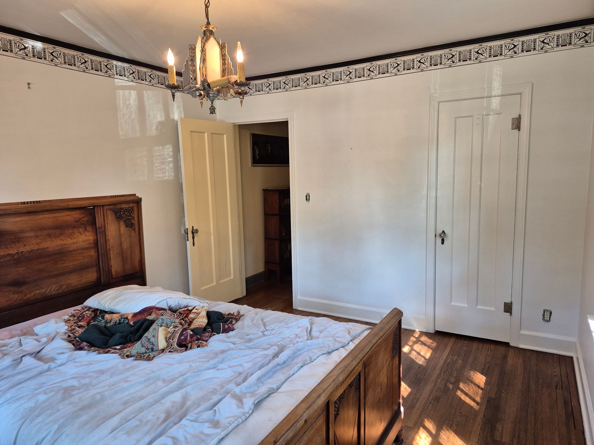 Bedroom with wooden bed frame, white walls, ornate chandelier, and open doorway to another room.