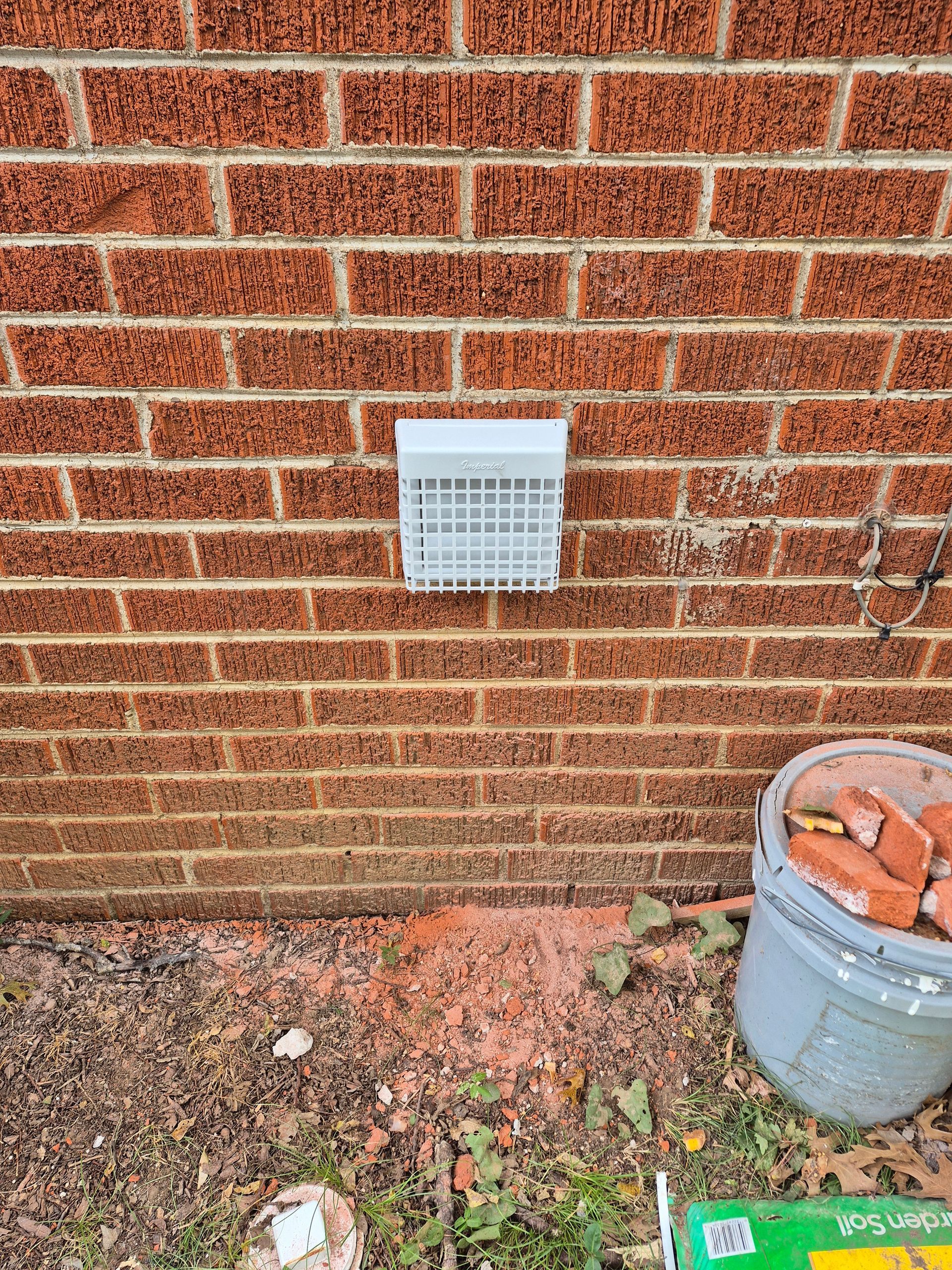 Brick wall with white vent, ground debris, and bucket of bricks.