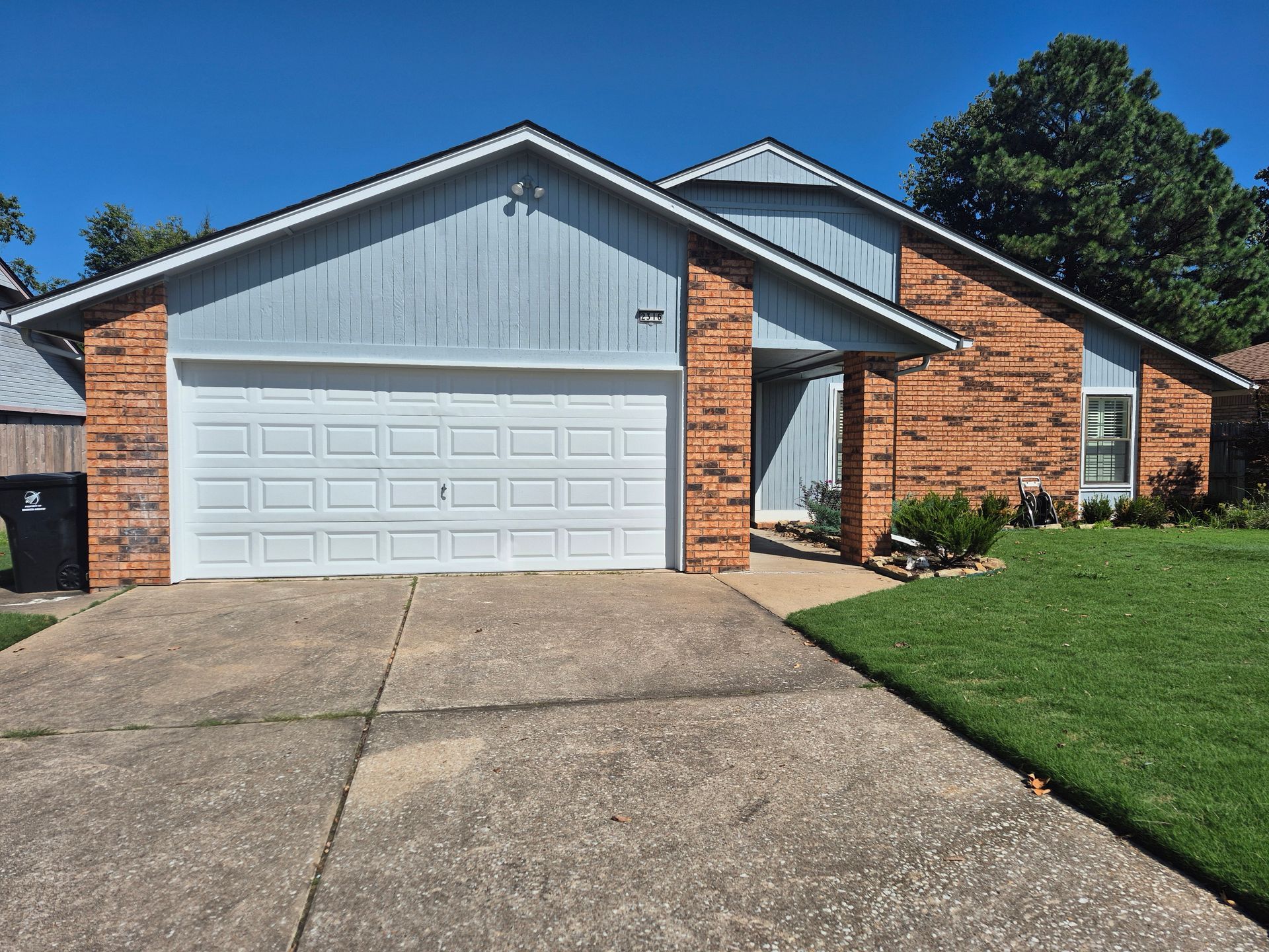 Blue-sided house with a white garage door, red brick facade, and a concrete driveway on a sunny day.