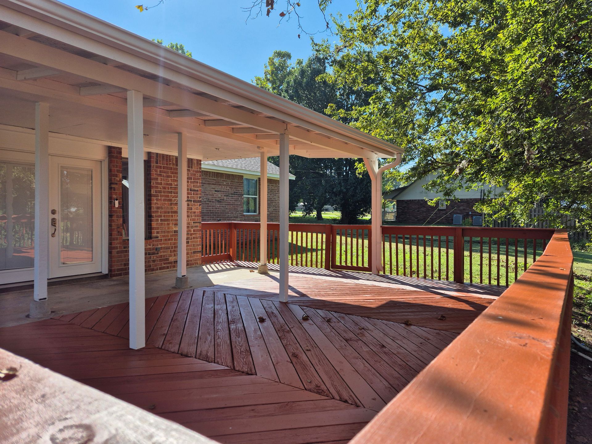 Covered wooden deck with railing, connected to a brick building. Bright day.