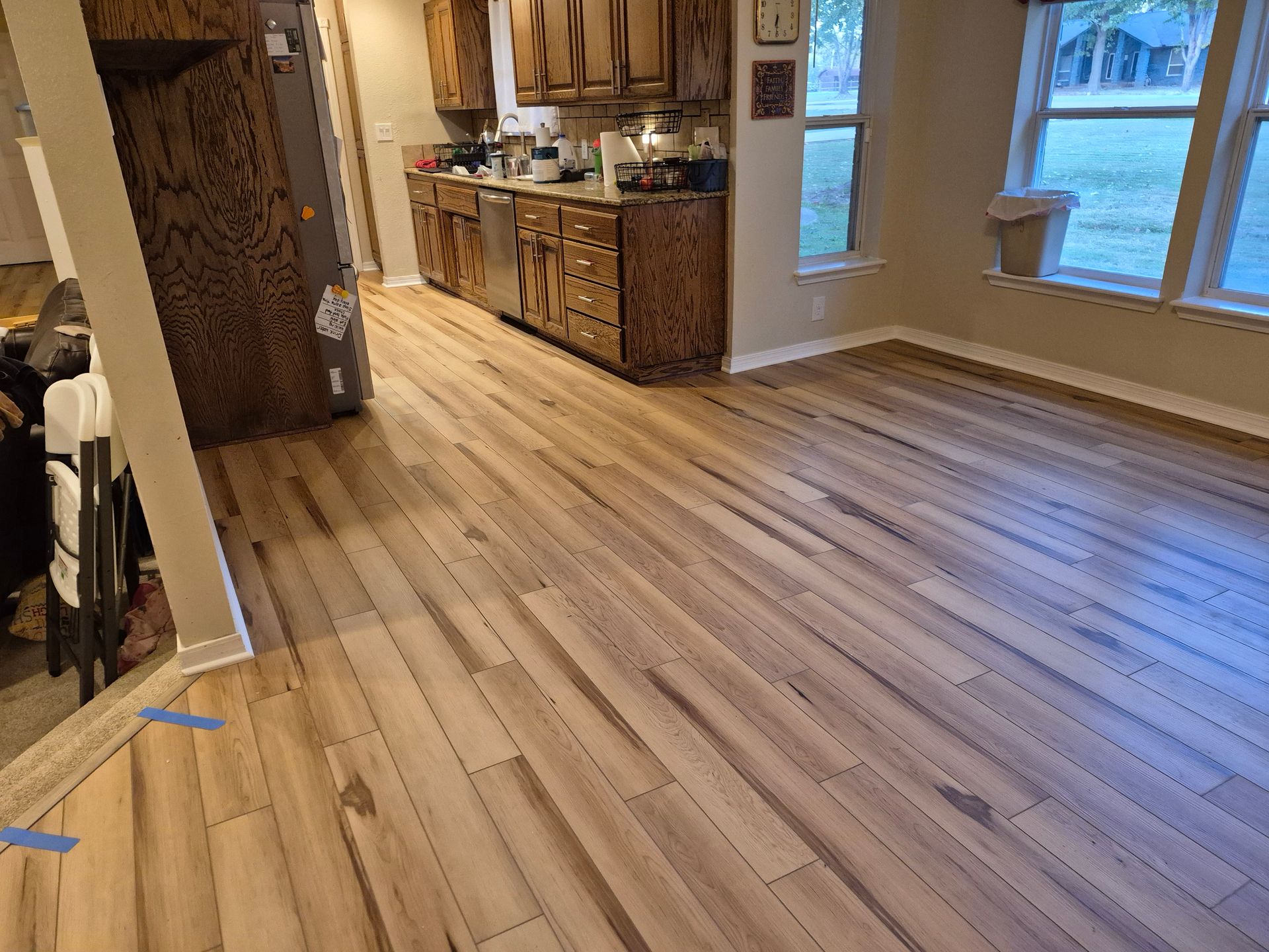 Light wood-look flooring extends from the kitchen into a living area with windows. Brown cabinets and a stainless steel appliance are visible.