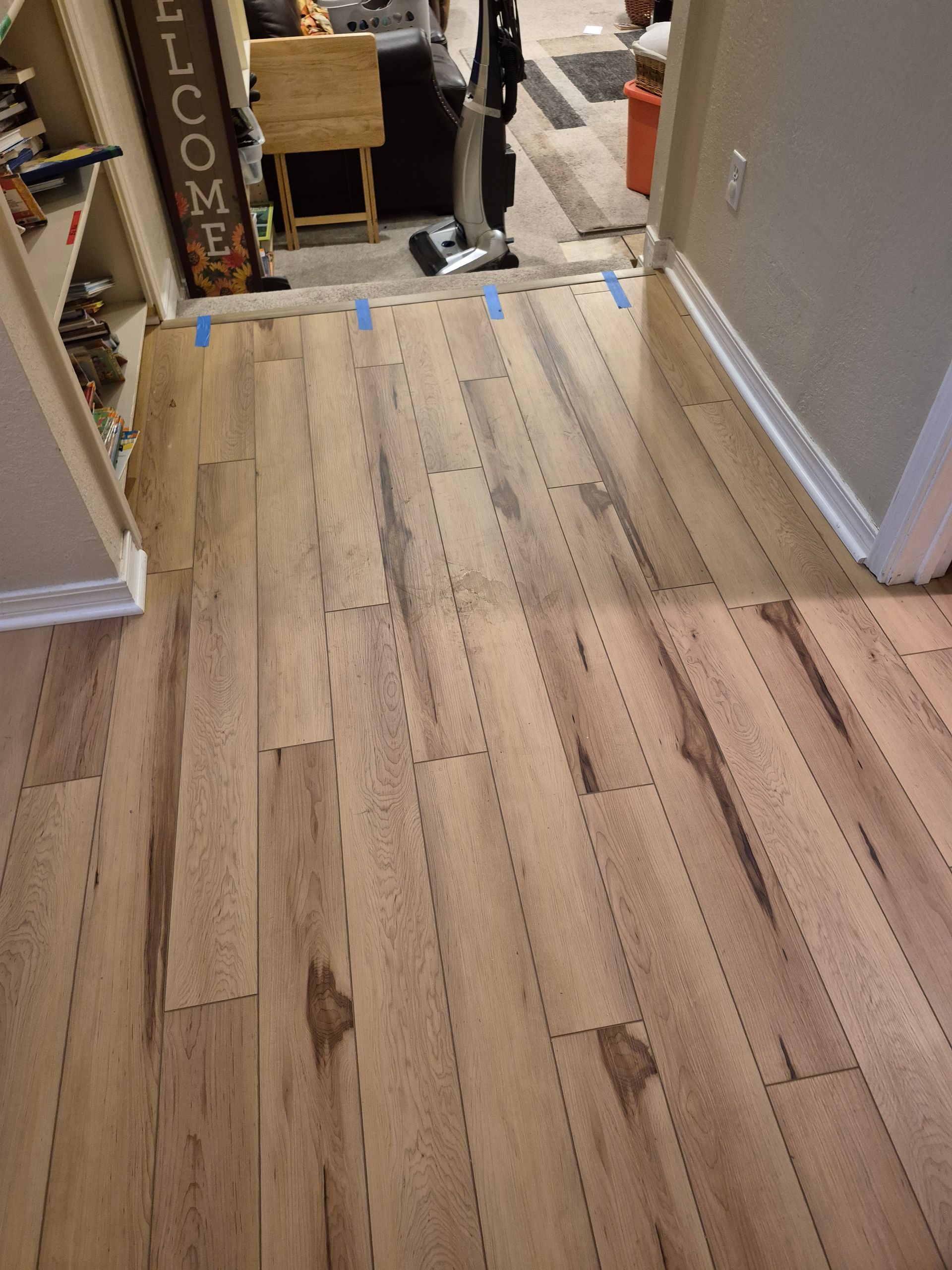 Wooden floor planks with cut lines, blue tape, and a hallway leading to a room with a vacuum.