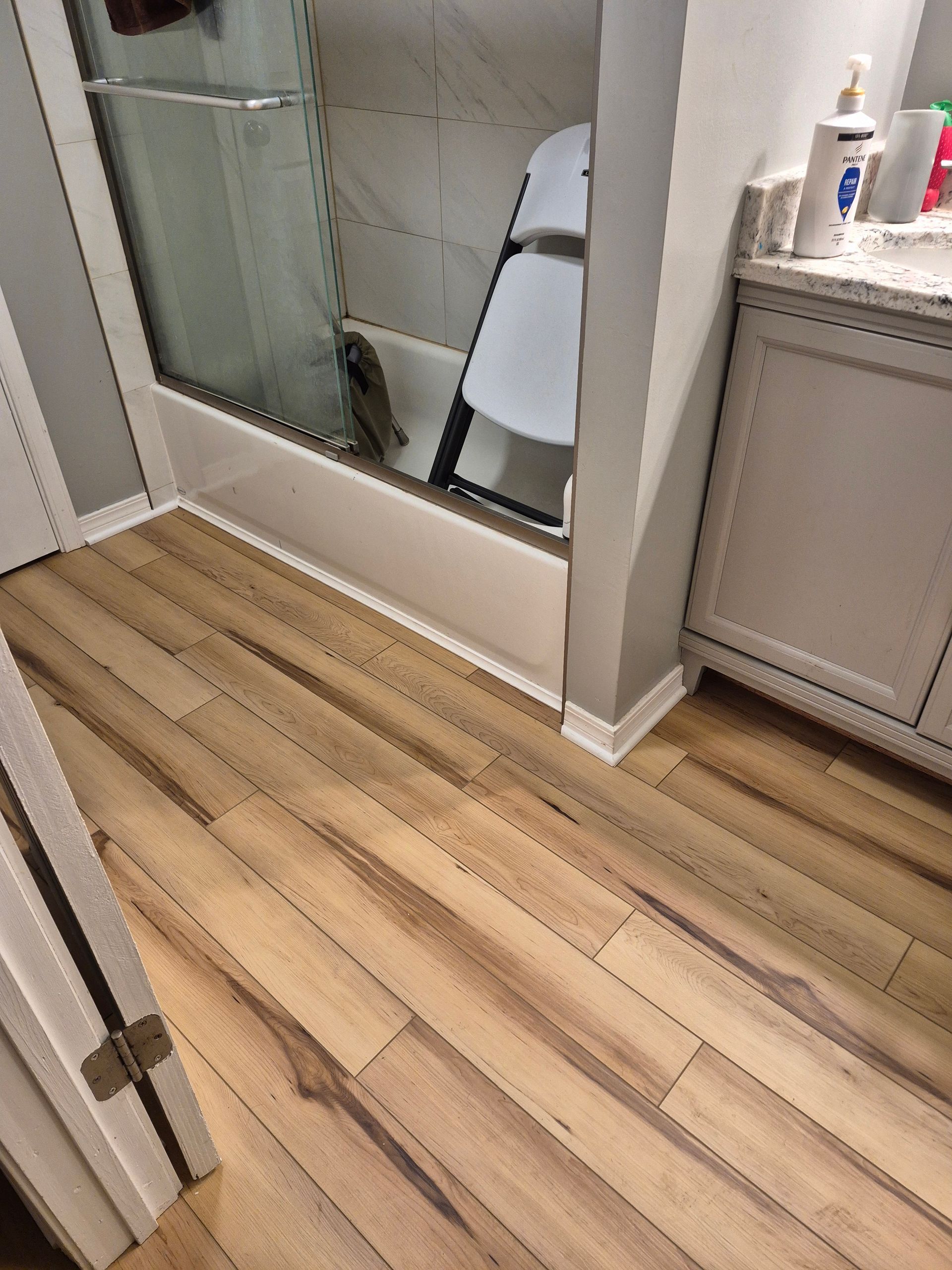 Bathroom with light wood-look flooring, a glass shower, gray vanity, and a folding chair inside the shower.
