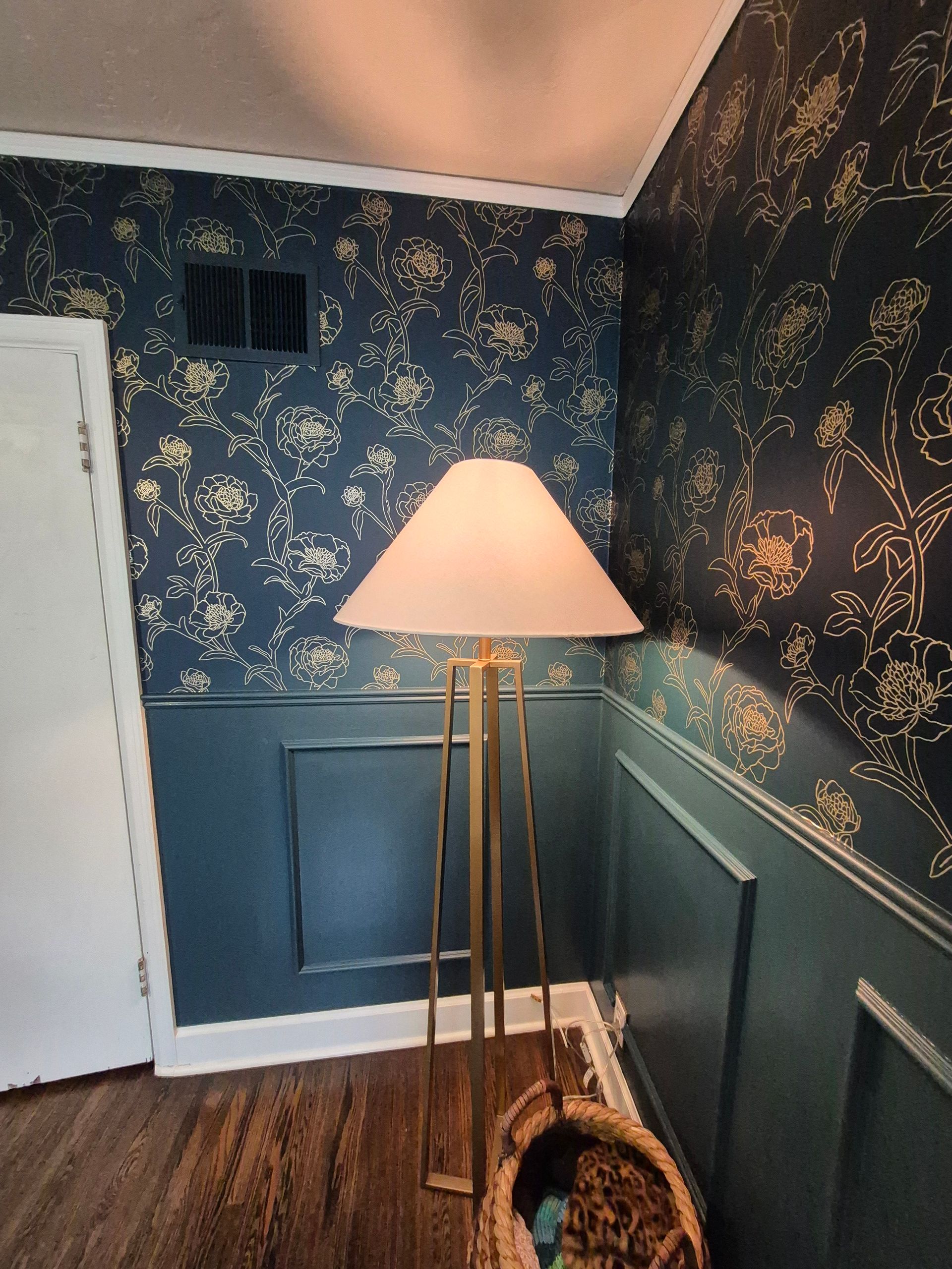 Gold floor lamp with a white lampshade stands near a patterned wallpapered wall.
