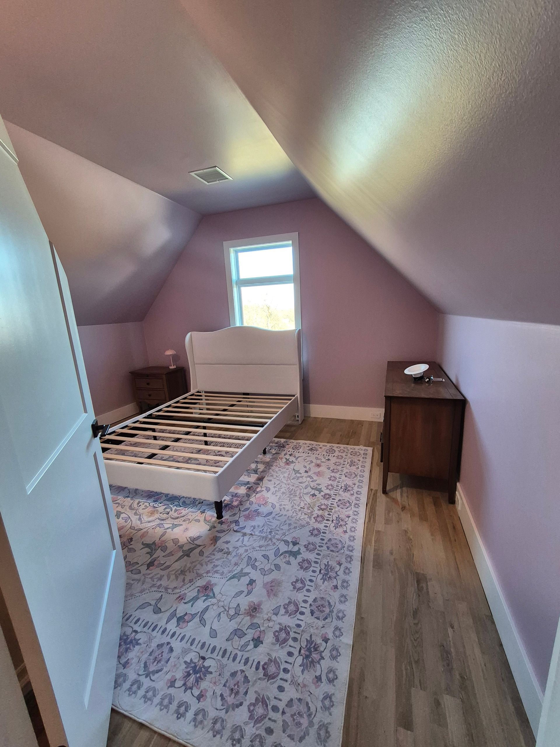 Bedroom with lavender walls, angled ceiling, white bed frame, rug, nightstands, and small window.