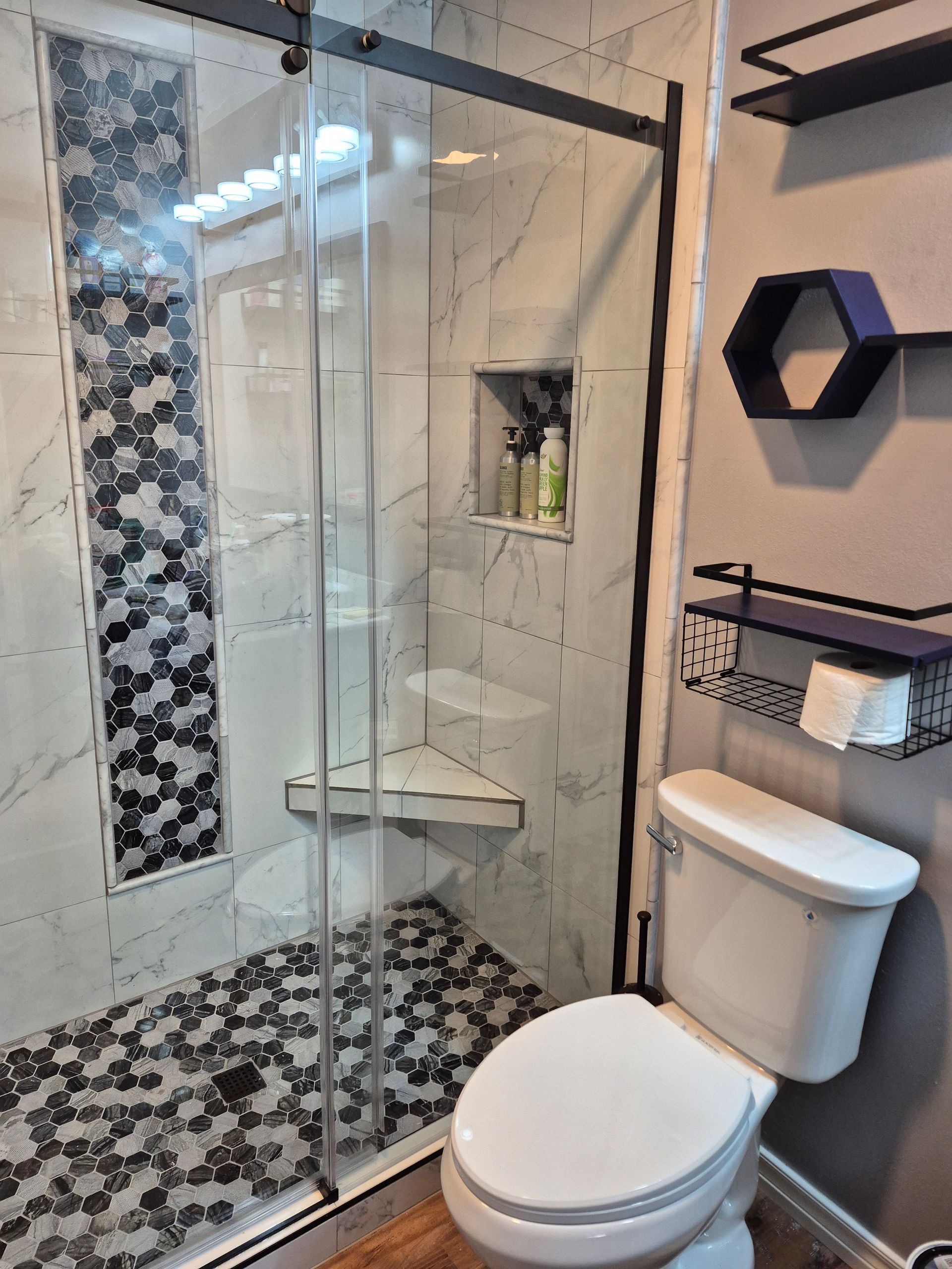 Bathroom with marble and hexagon tile shower, glass door, corner seat, and white toilet.