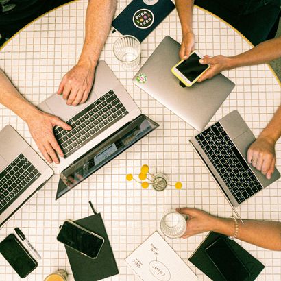 A group of people are sitting around a table with laptops and phones.