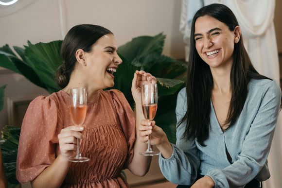 Two people laughing while holding glasses of pink sparkling wine, seated in front of large green tropical leaves.