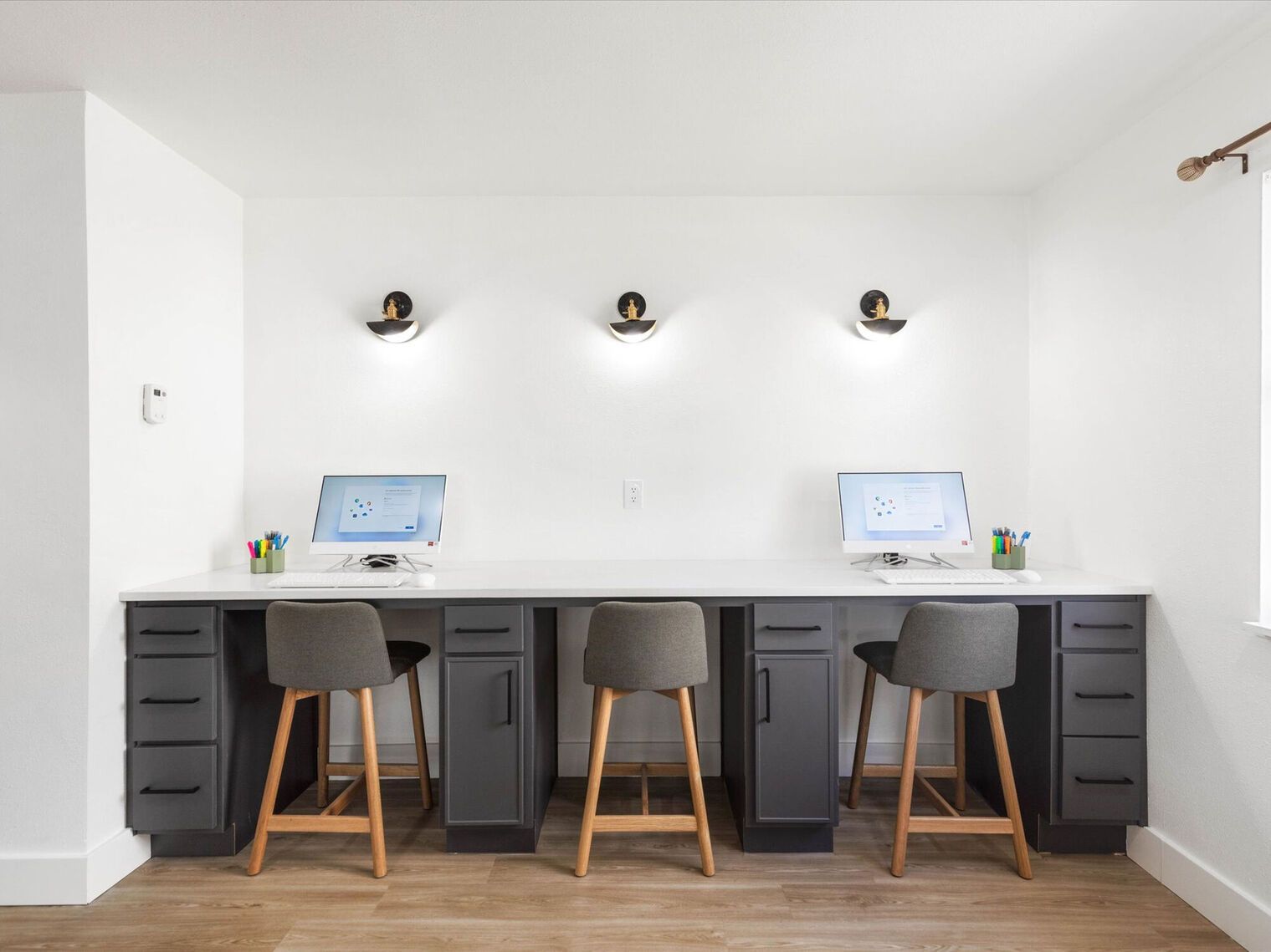 Three workstations with computers, gray cabinets, gray stools, and three sconces against a white wall.