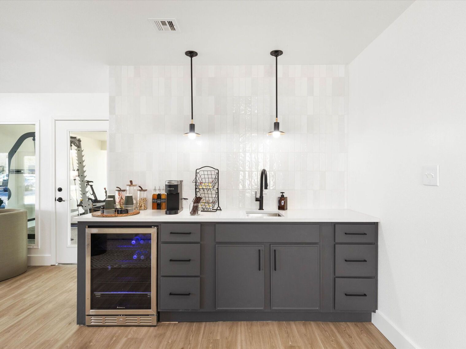 Gray cabinet bar with a wine cooler, sink, white countertop, and pendant lights.