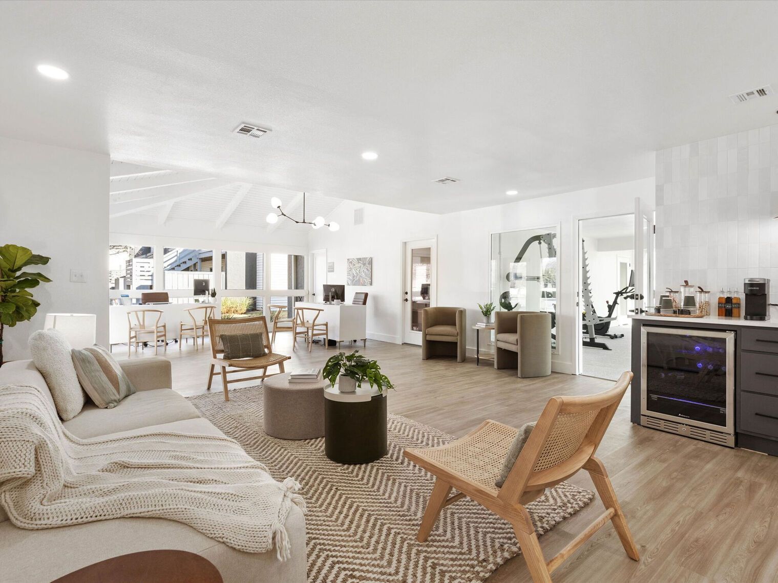 Spacious white living room with light wooden floors, seating, and a view into a kitchen and home gym.