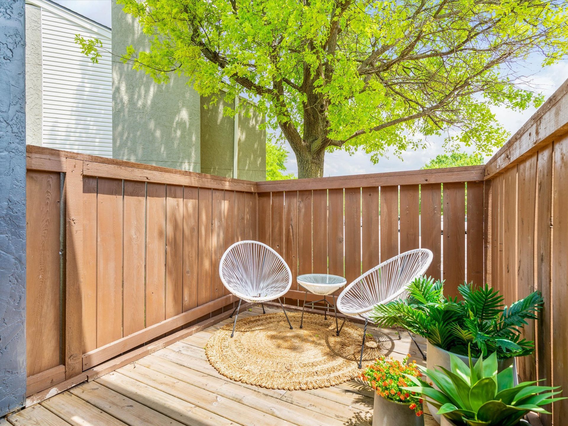 Cozy outdoor patio with two white woven chairs, a small table, a round rug, and potted plants.