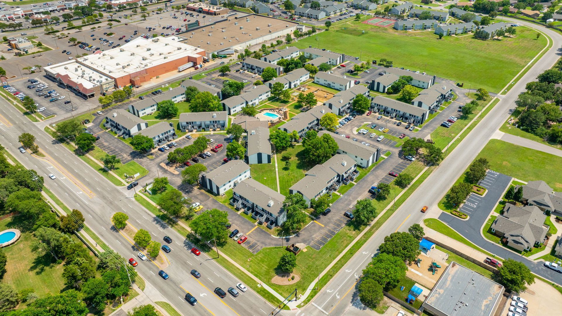 Aerial view of a residential complex with multiple buildings, a pool, and a shopping center nearby.