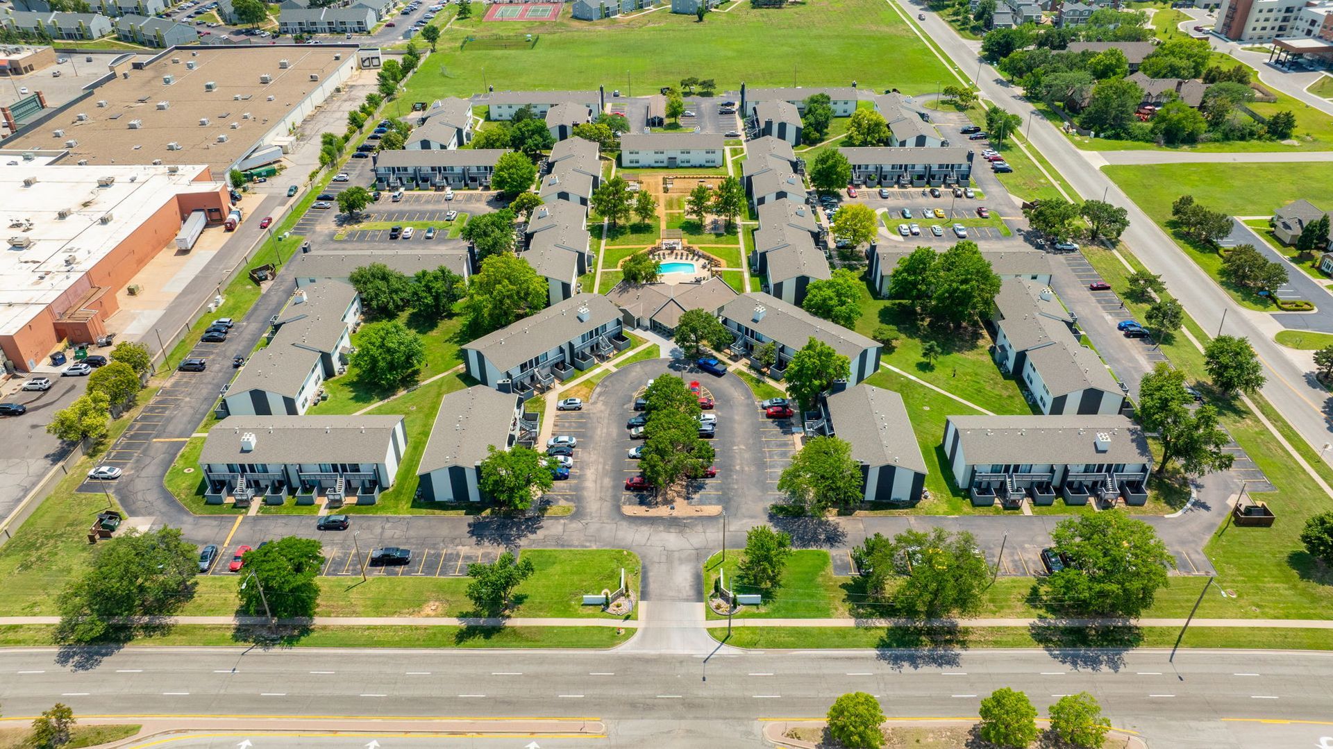 Aerial view of a residential complex with multiple apartment buildings, green lawns, trees, and a central pool.