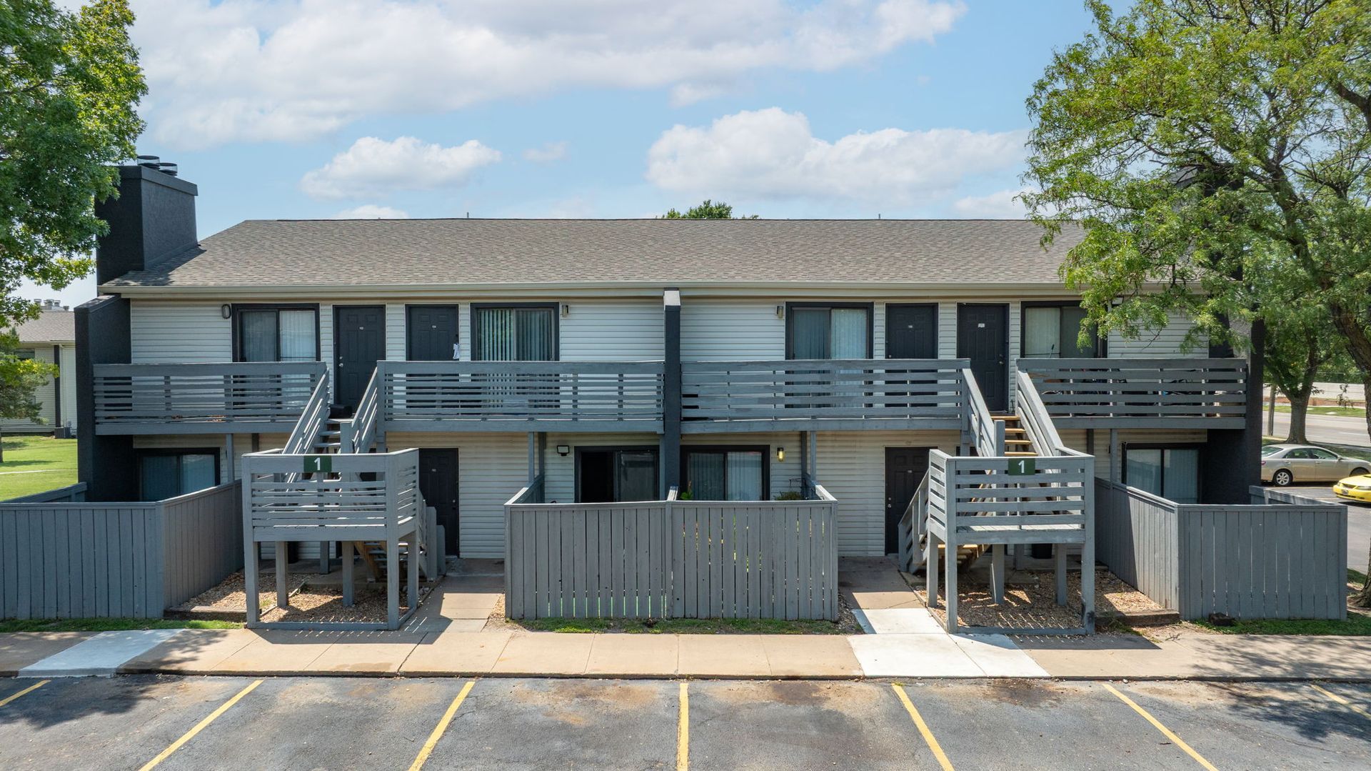 Two-story apartment building with gray siding, balconies, stairs, and a parking lot. Blue sky.