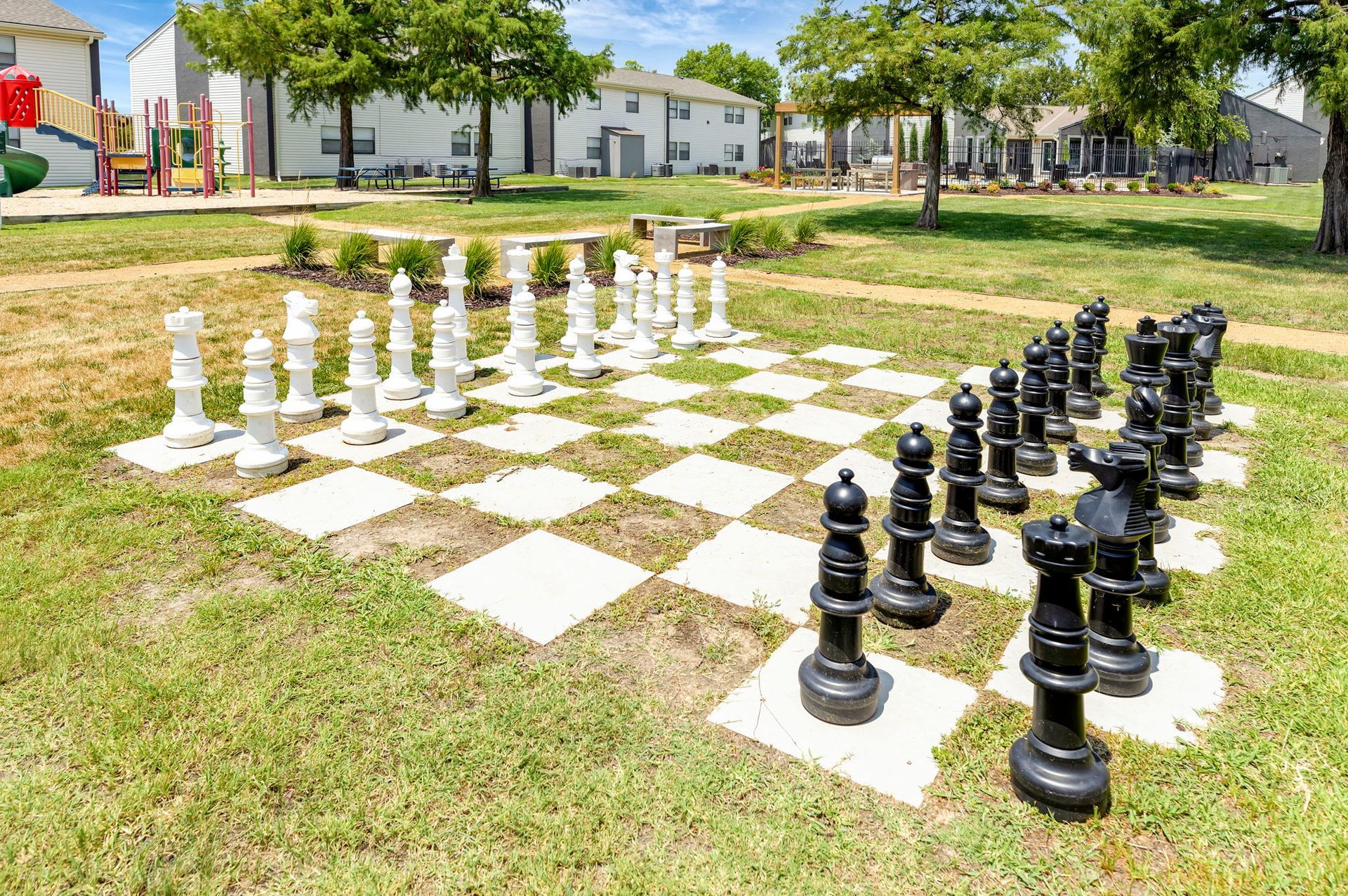 Large outdoor chessboard with black and white pieces on a grassy area near townhouses.