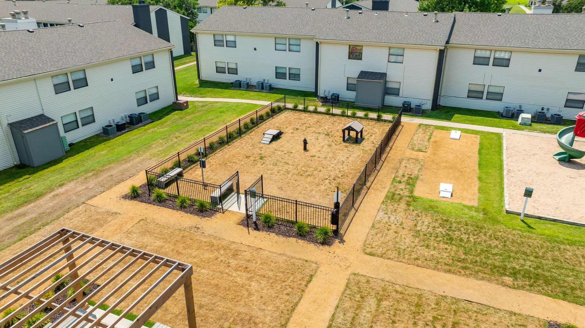 Aerial view of a fenced dog park with play structures, seating, and a pergola near apartment buildings.