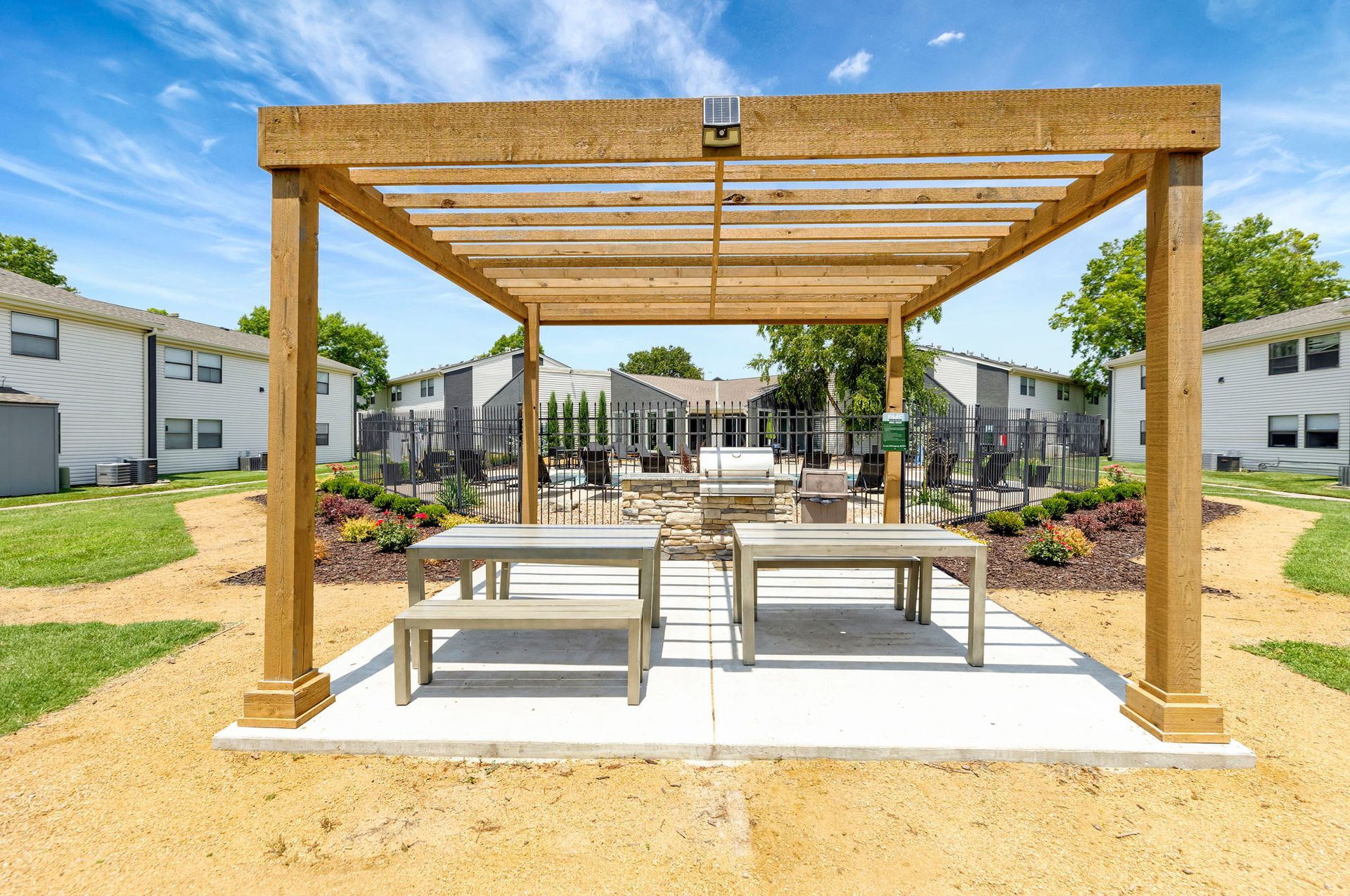 Wooden pergola with benches over a concrete patio; background shows outdoor seating area and white buildings.