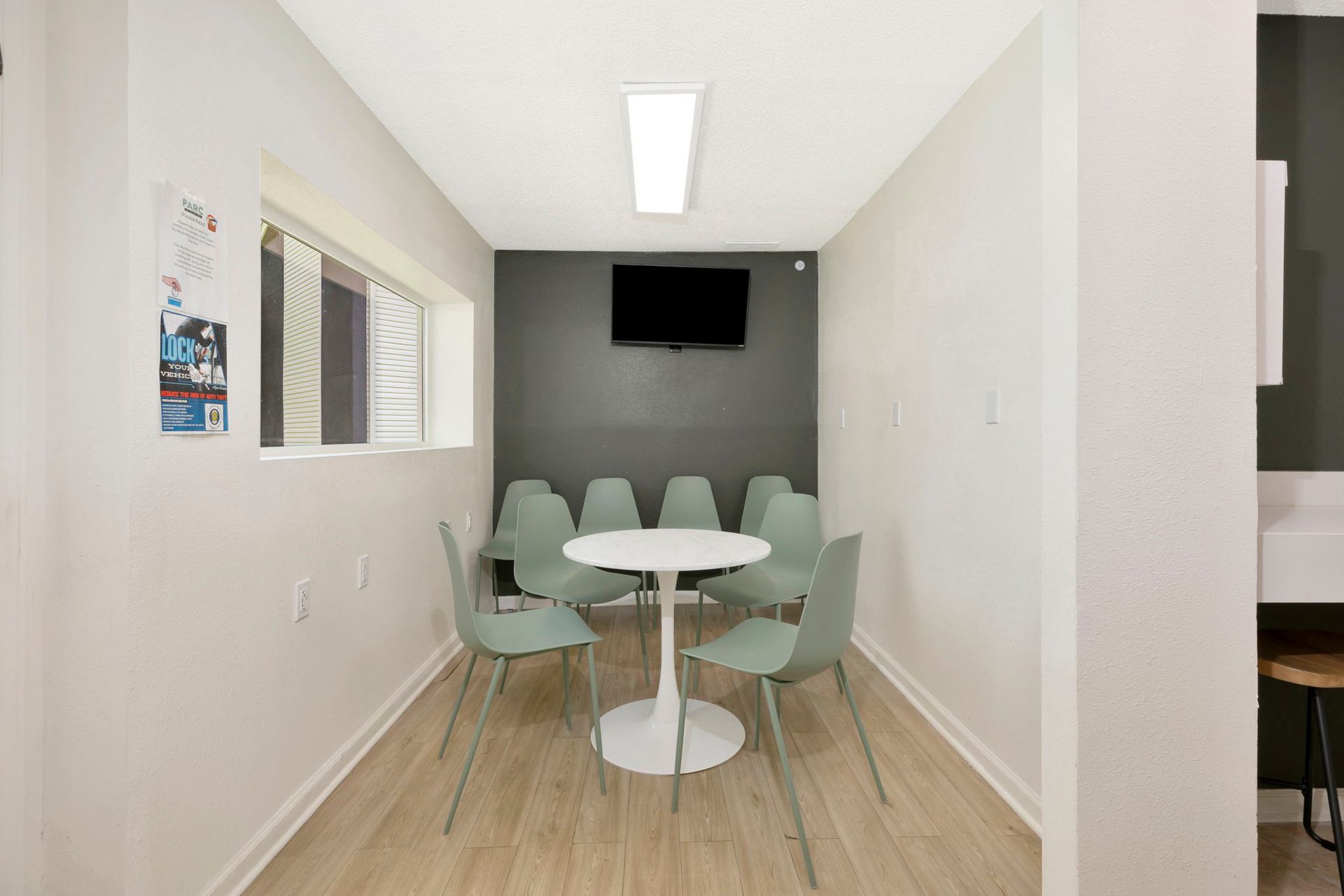 Narrow room with round table, eight green chairs, TV on dark gray wall, and window.