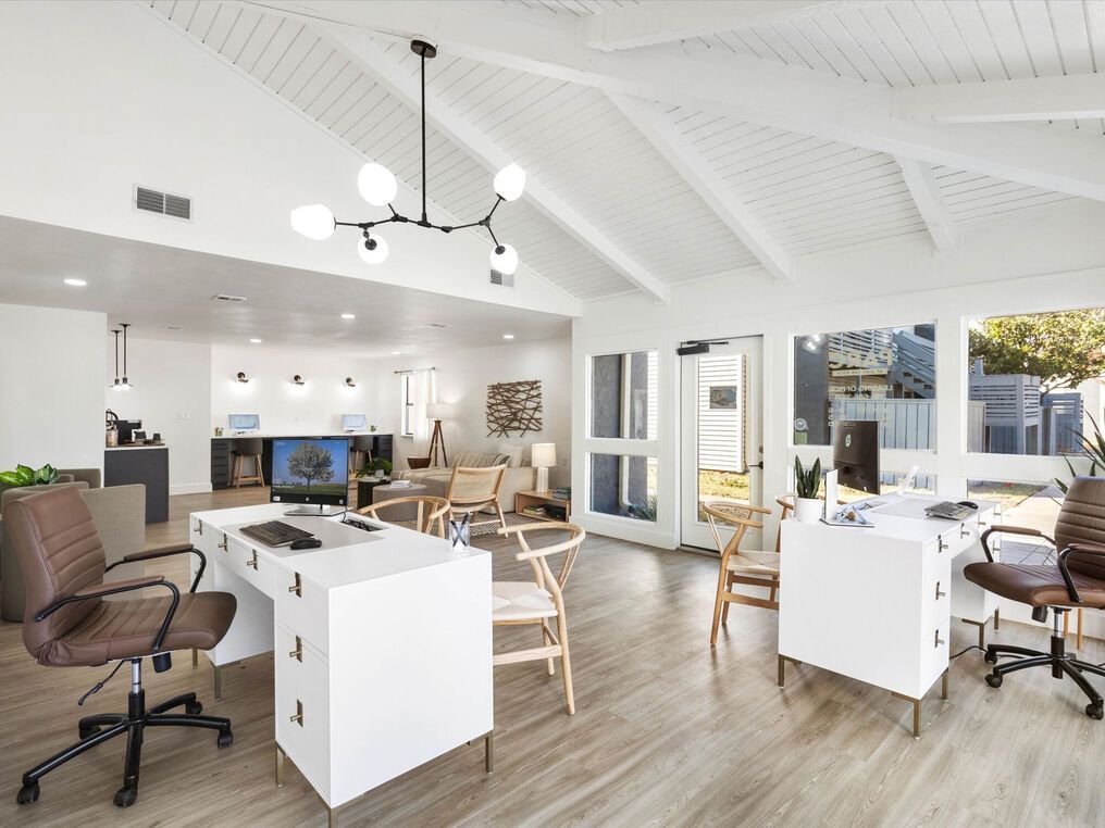 Bright office interior with desks, chairs, and natural light from large windows. Ceiling beams and modern light fixture.