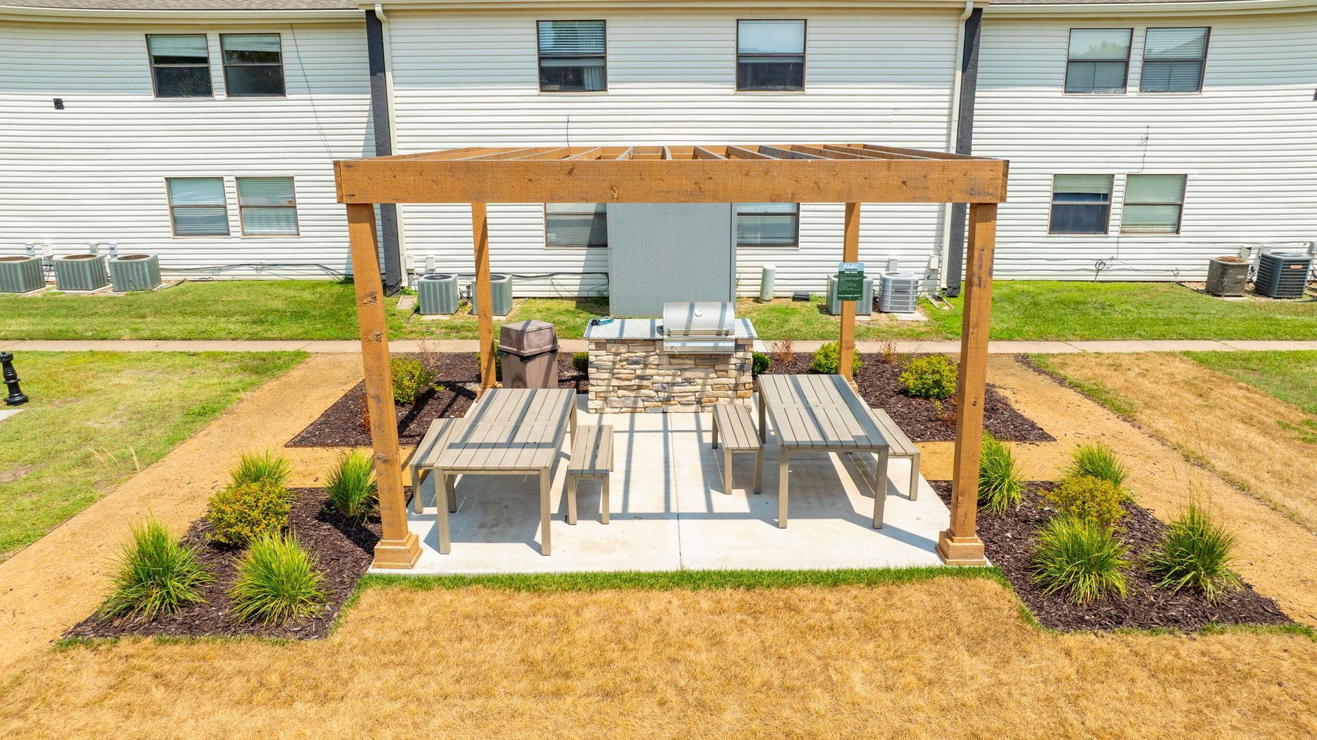 Outdoor grilling area with wooden pergola, stone grill, and two picnic tables.