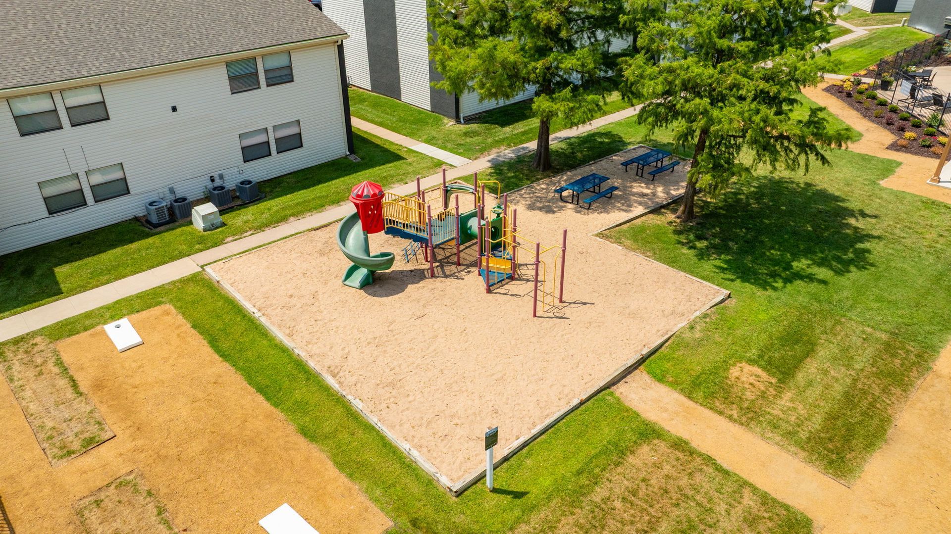 Playground with slide, climbing structures, and picnic tables in a grassy area beside a building.